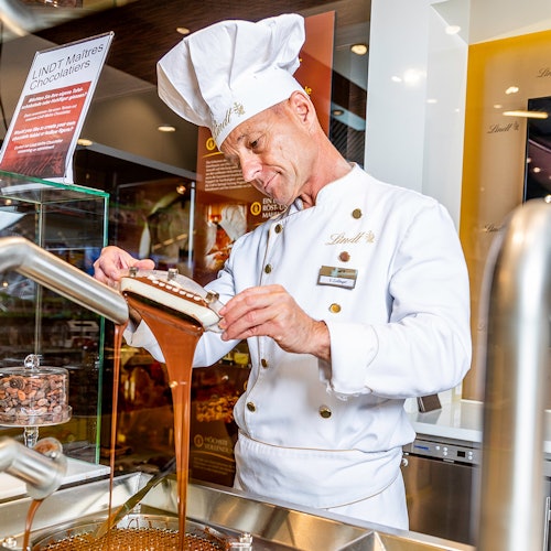 A chocolatier in a white uniform and hat, pouring liquid chocolate from a mold in a Lindt shop.