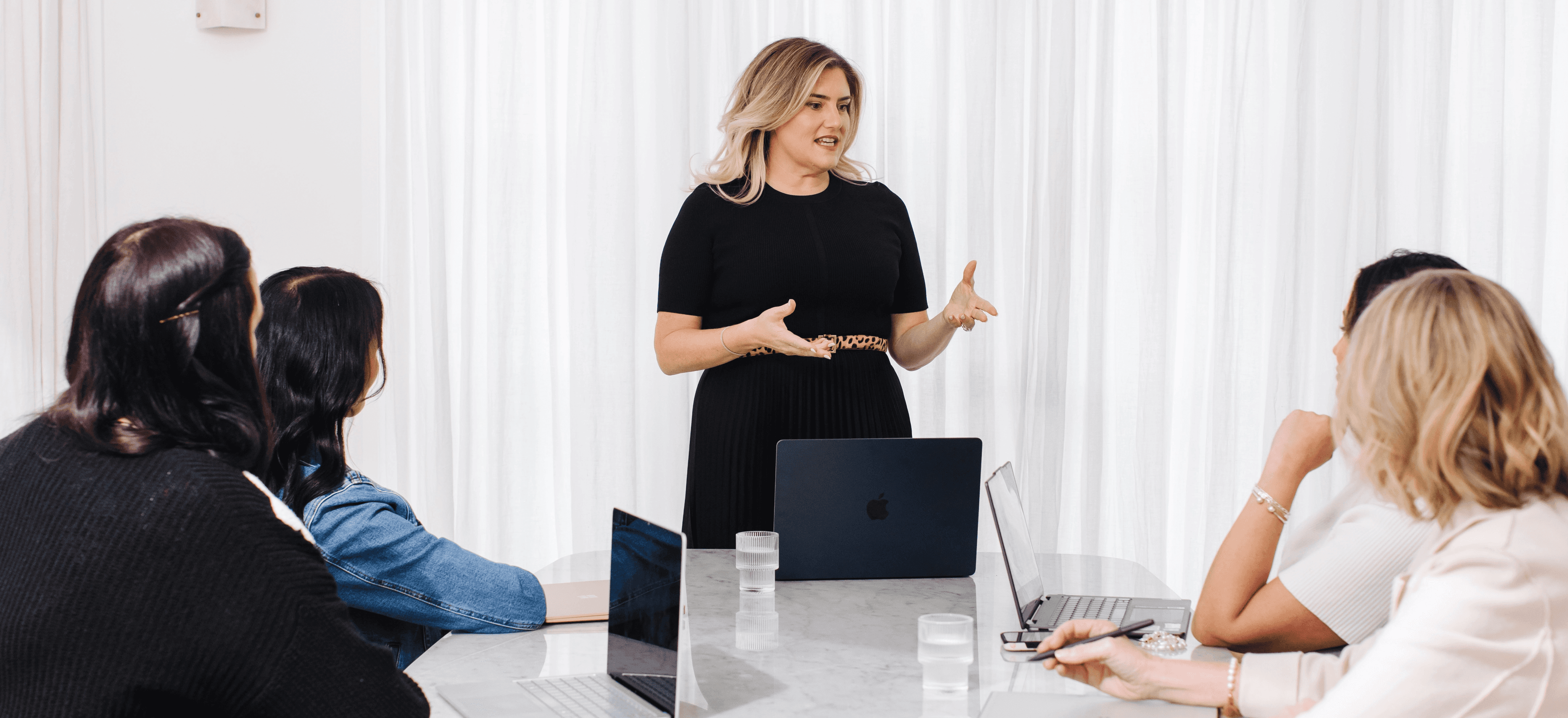 Woman in a black dress standing, speaking to four women seated at an table with laptops open.