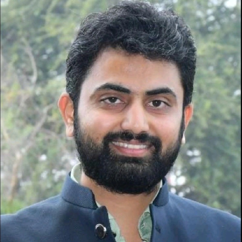 A middle-aged man with gray hair and glasses, wearing a dark apron, standing and looking directly at the camera with a neutral expression against a blurred, metallic kitchen background.