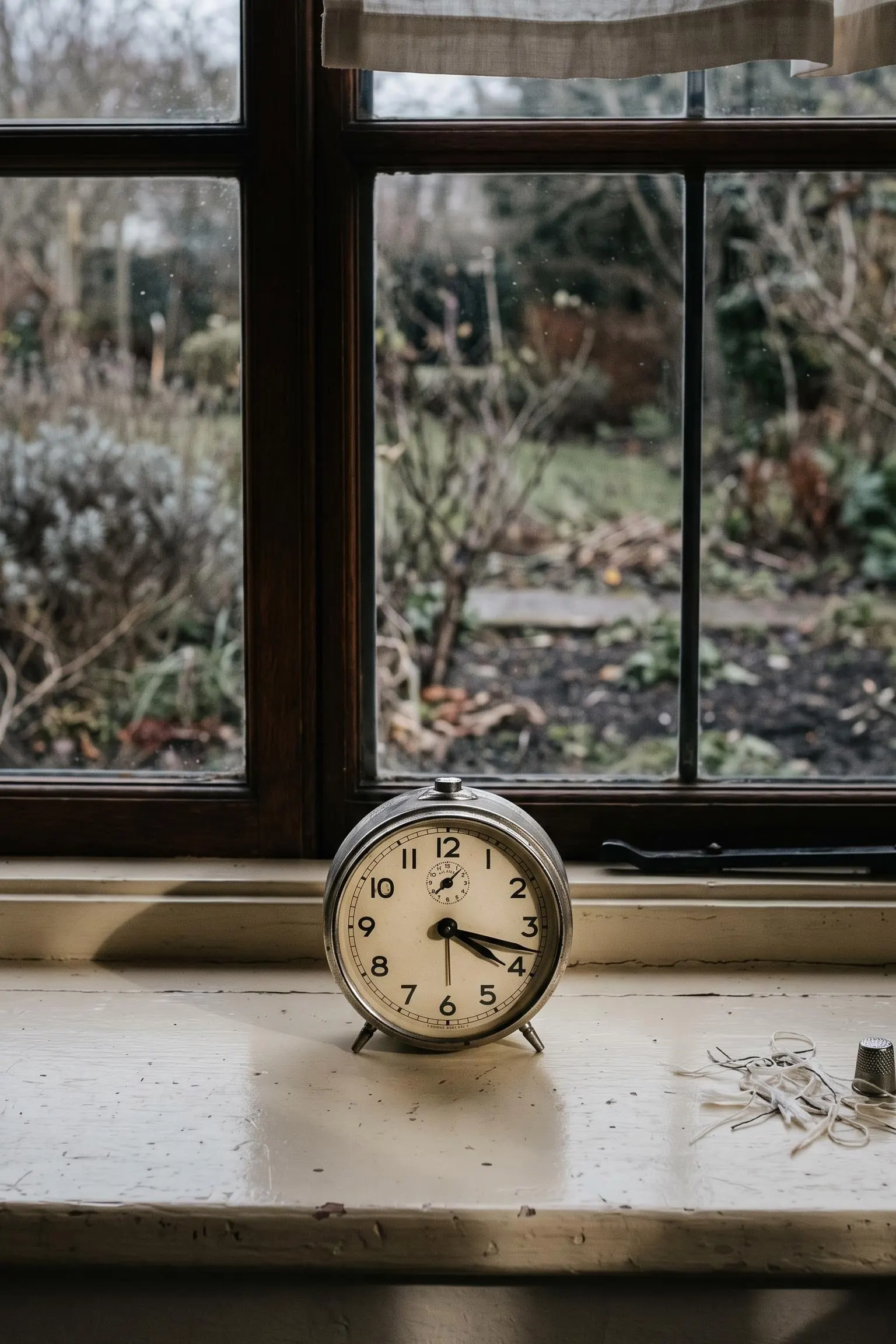 A wind-up alarm clock on a painted windowsill in low winter light, long shadows across the paint