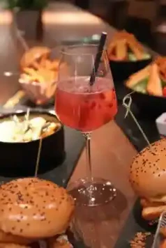 Roter Cocktail im Weinglas, serviert auf einem Tisch mit Burgern und verschiedenen Gerichten im kAlps.