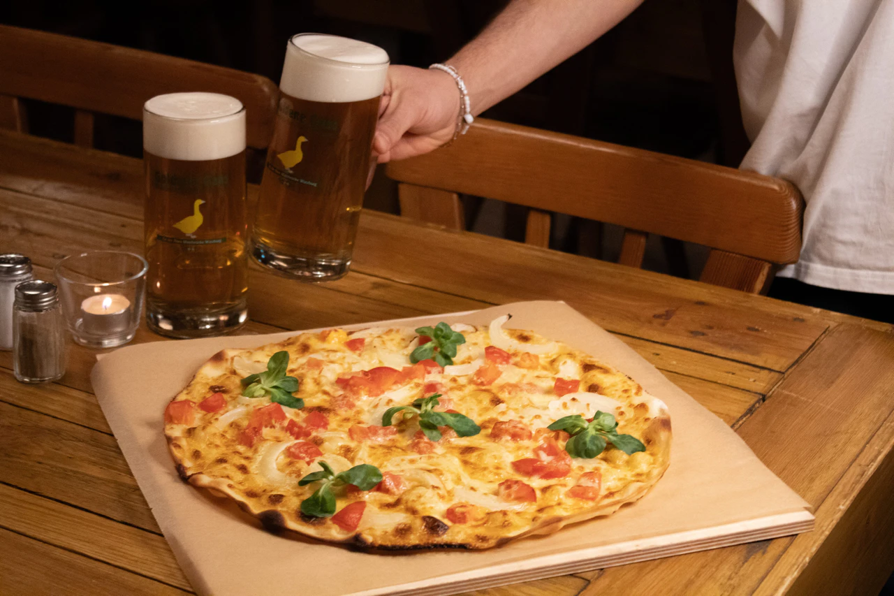 Flammkuchen auf einem Tisch mit zwei Biergläsern; eine Hand greift nach einem Glas.