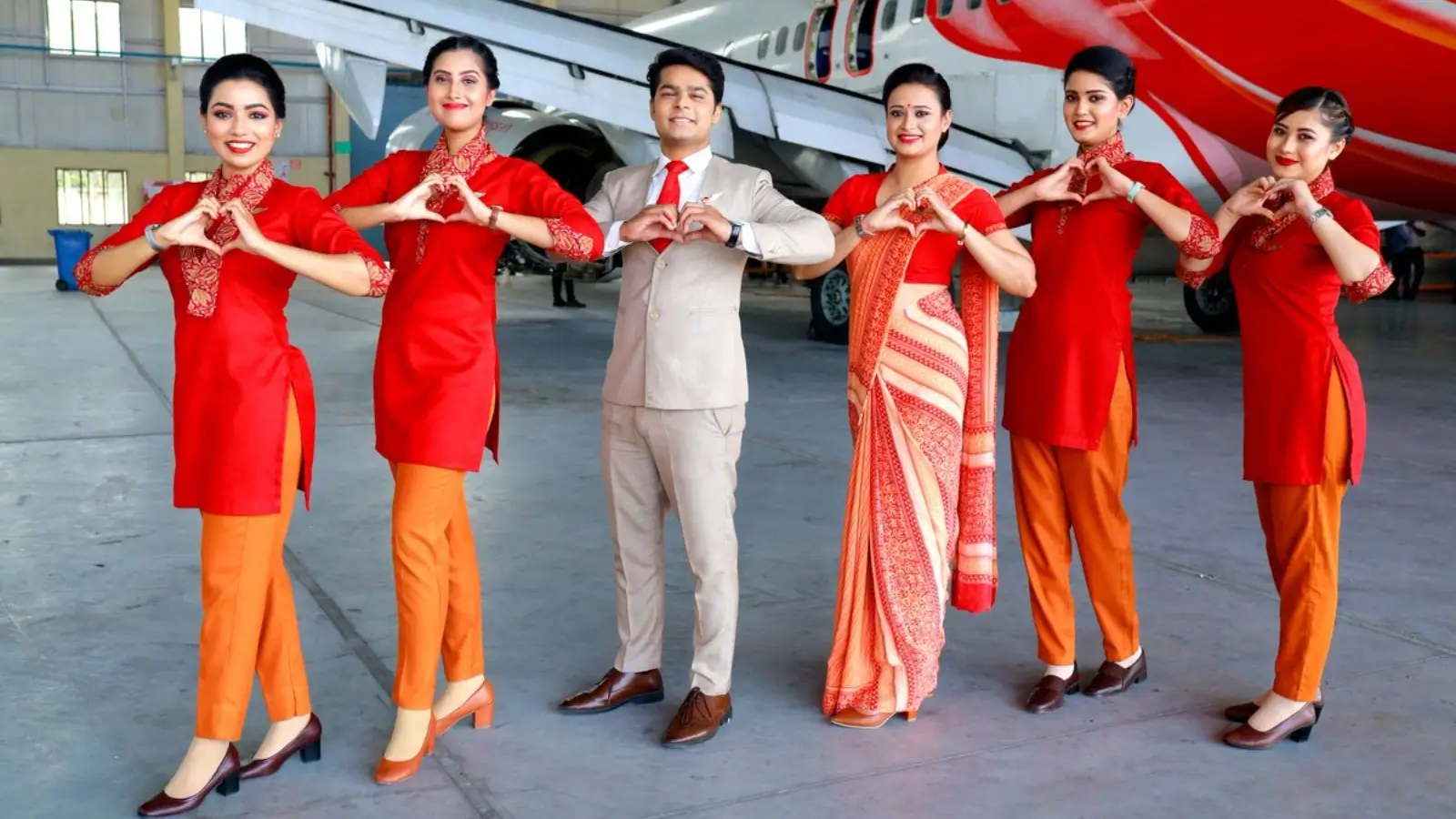 Six Air India cabin crew members walking confidently on an airport tarmac in front of a large jet engine. The women are wearing elegant red and purple sarees and tunics, while the men are in sophisticated high-neck "Bandhgala" suits in burgundy and black.