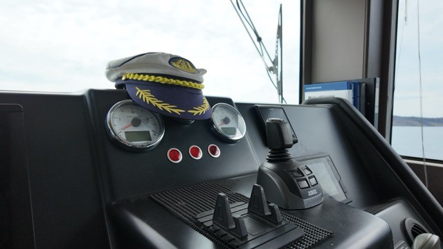 Cranchi T55 Eco Trawler flybridge helm station interior showing the dark dashboard with analogue speed and engine gauges, red warning buttons, joystick docking control, throttle levers, and a captain's peaked cap resting on the console, with panoramic windscreen views over open water