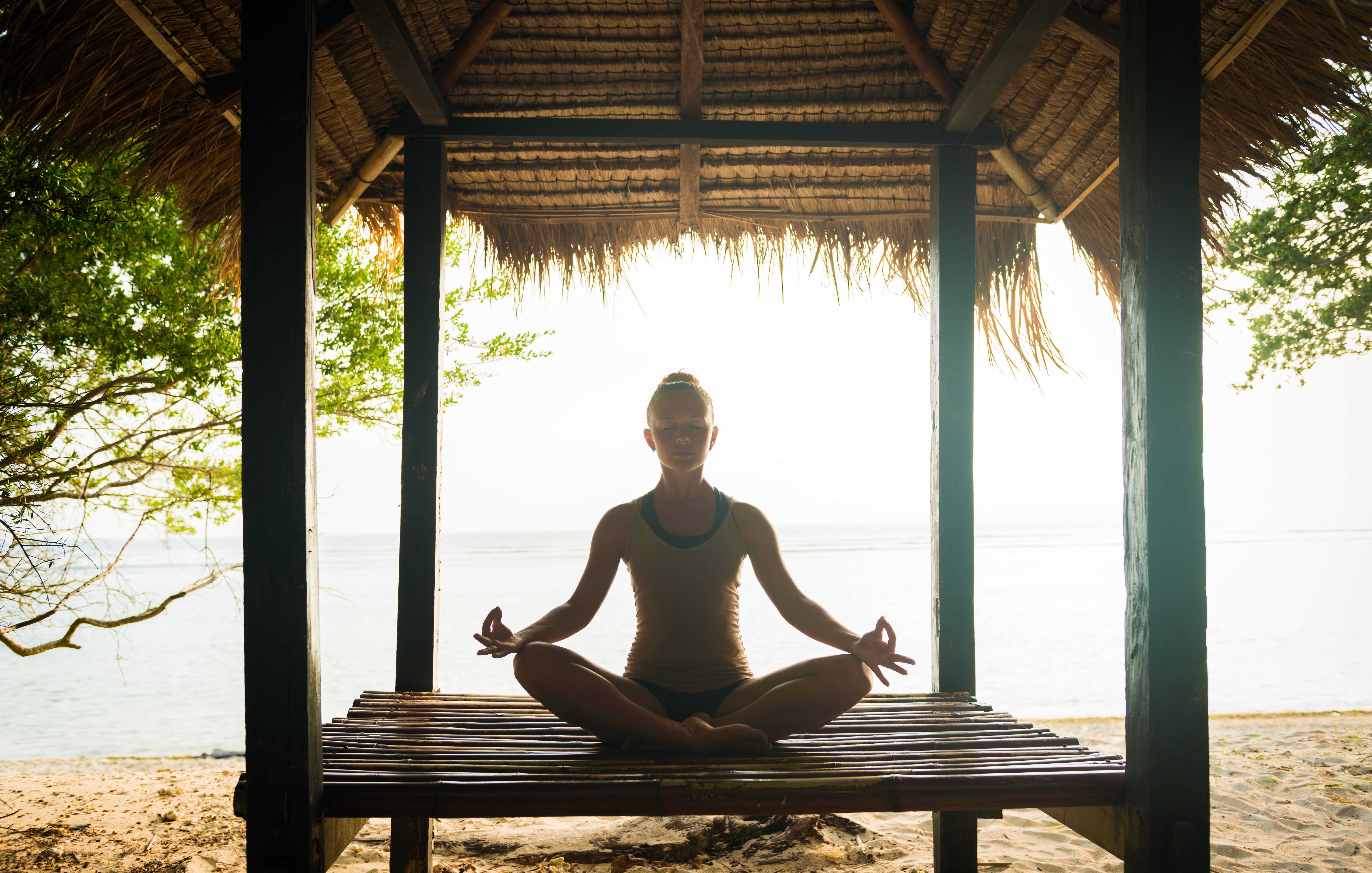 A circle of people sitting cross-legged, meditating with hands in a prayer position. Soft, warm lighting.