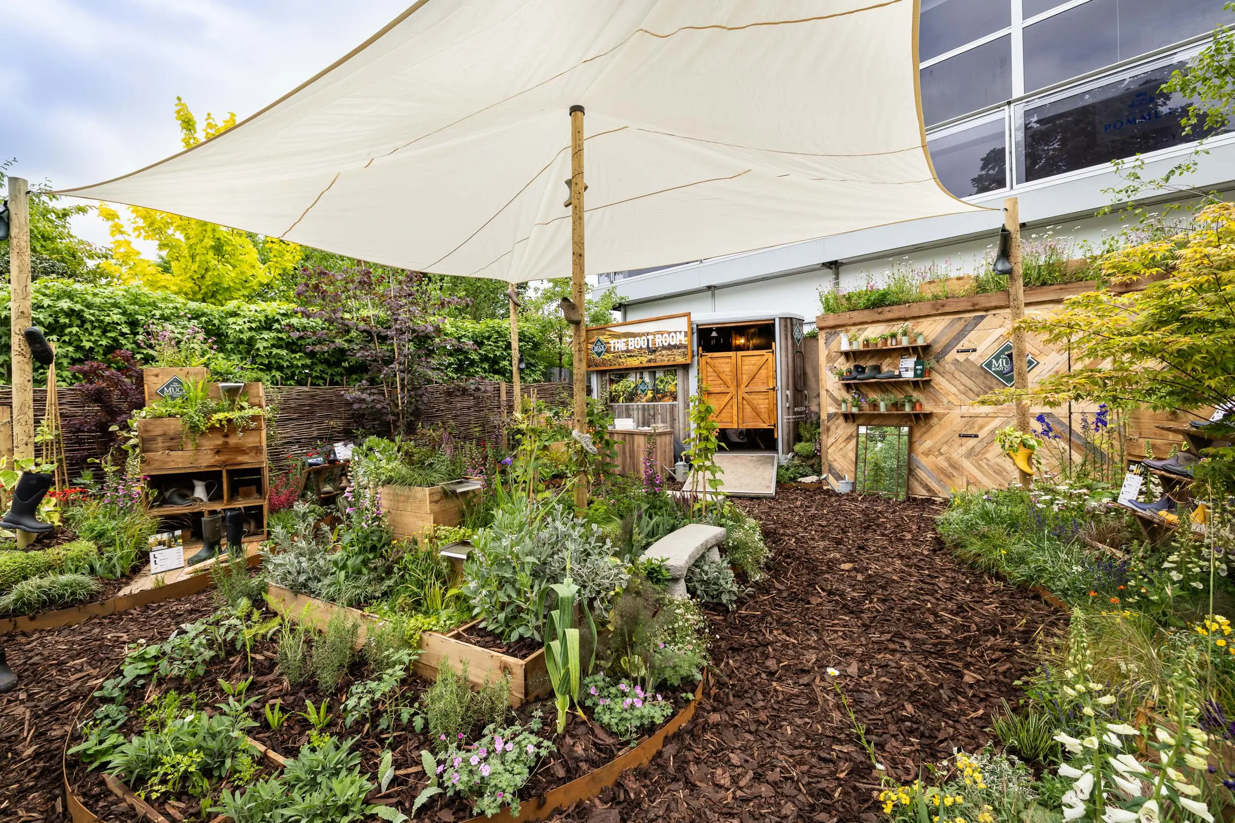 A lush garden with a variety of plants under a shade structure, featuring wooden pathways and vibrant greenery.