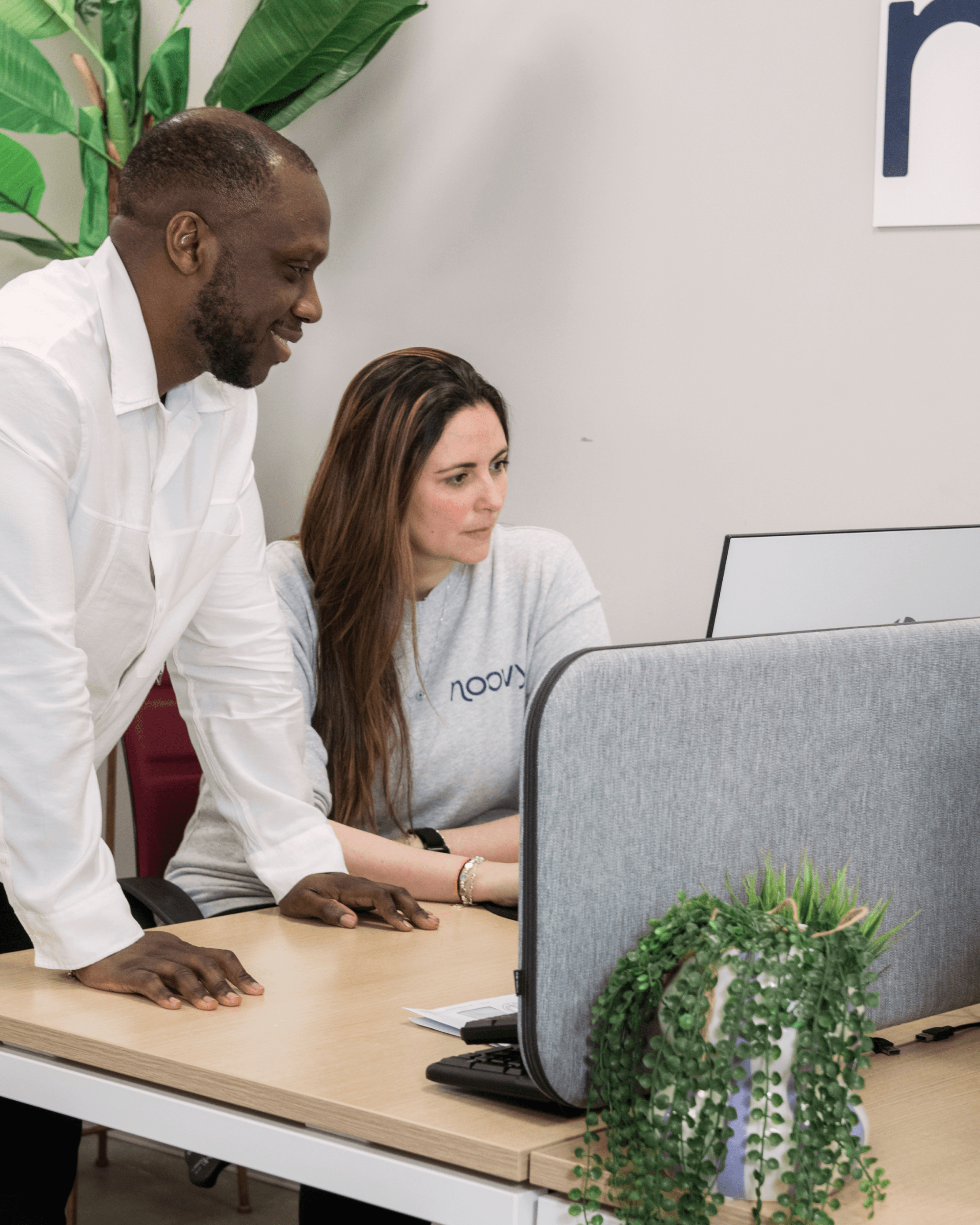 A man and a woman collaborate at a desk, focused on a computer screen, with plants in the background.