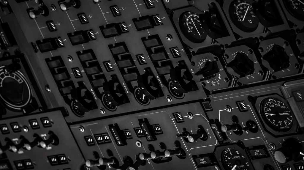 Black and white close-up of an aircraft control panel with switches, dials, and levers in a structured layout.