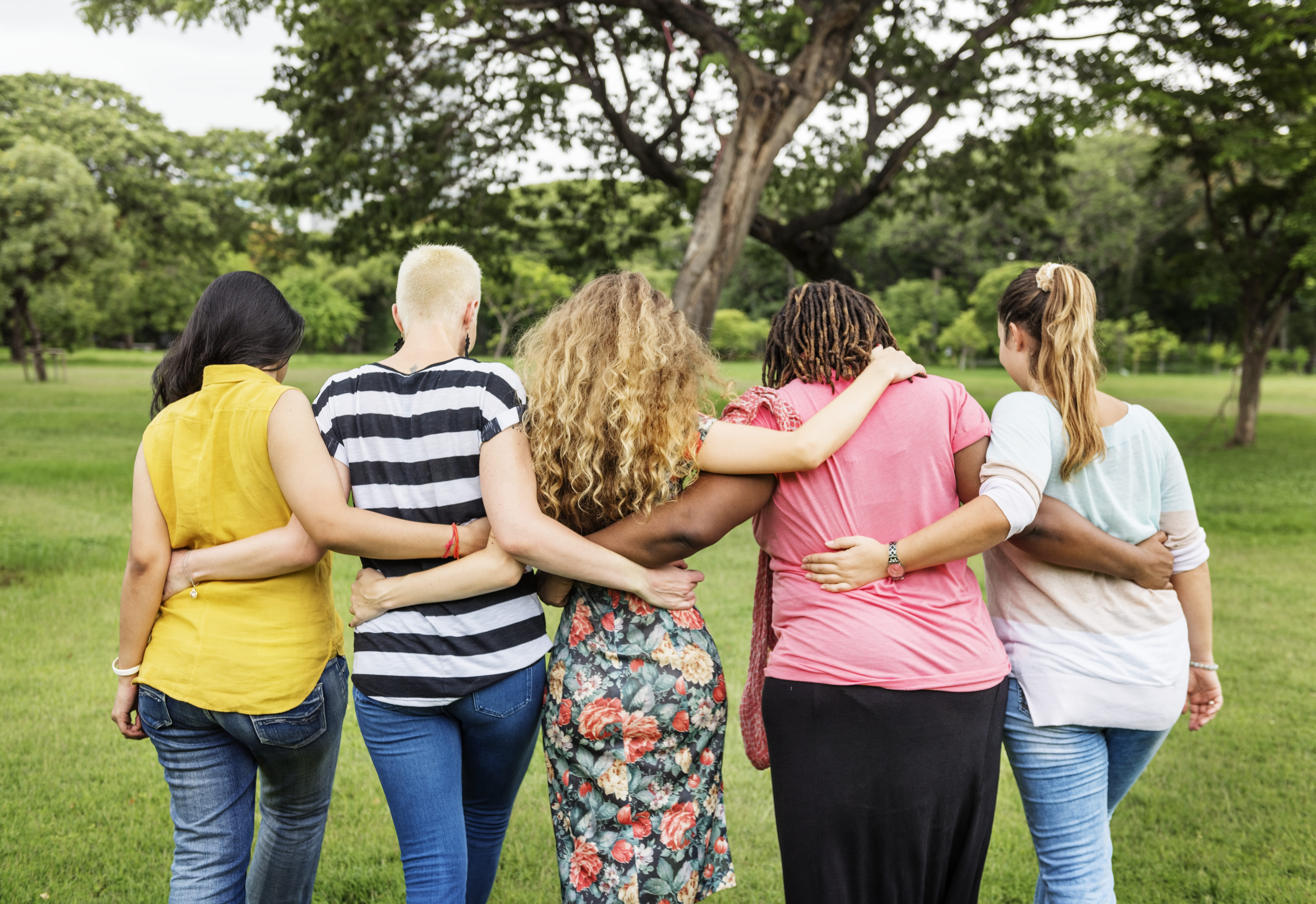 Imagem de um grupo de cinco mulheres andando juntas em um parque, de costas para a câmera. Elas caminham com os braços em volta uma das outras, demonstrando união e amizade. Cada uma veste roupas casuais e de estilos variados, refletindo diversidade e individualidade. A paisagem ao redor é verde e cheia de árvores, sugerindo um ambiente tranquilo e natural, ideal para momentos de confraternização e apoio mútuo.