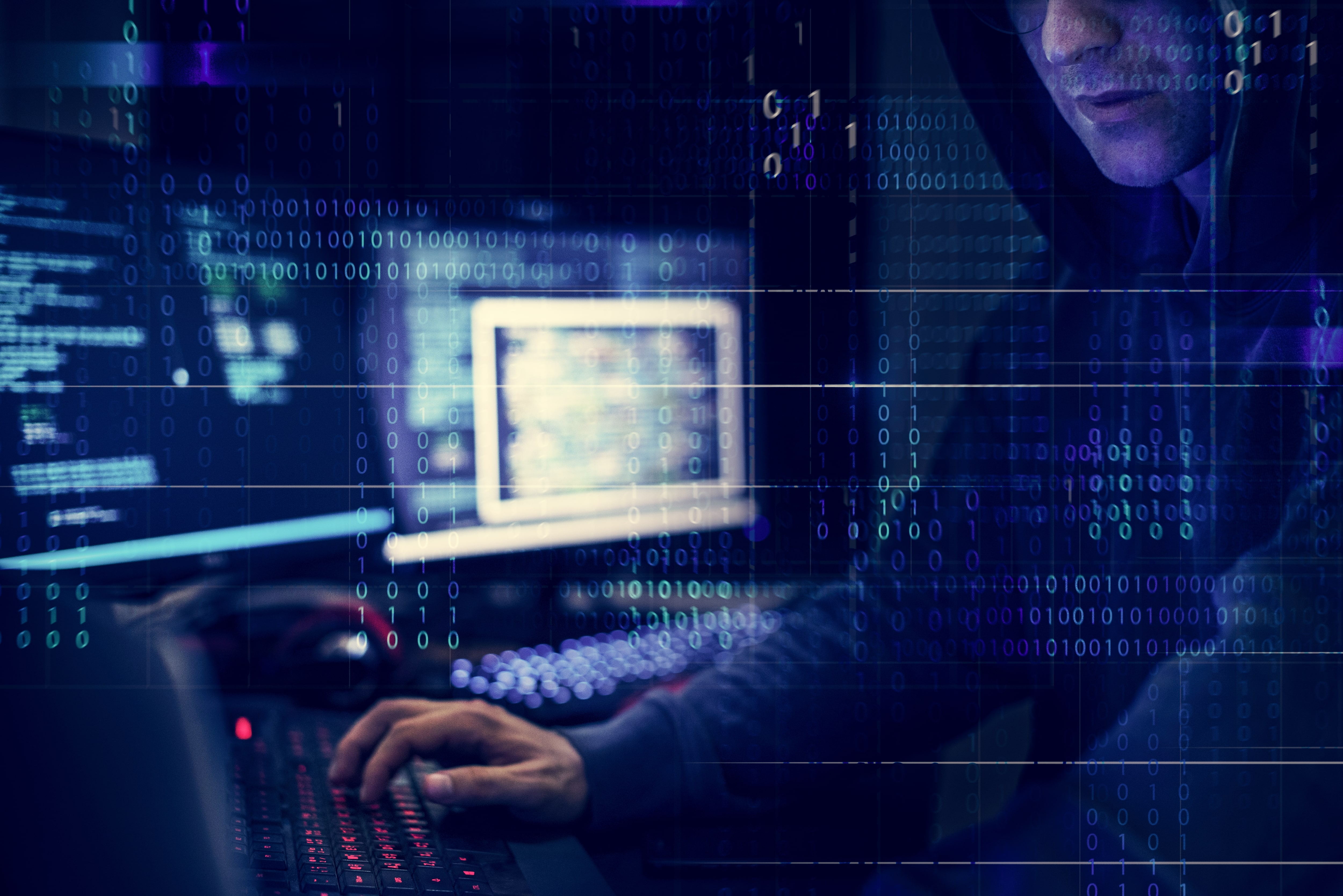 A hacker in a dark hoodie works at a multi-monitor computer setup in a dark room, with a digital overlay of streaming blue binary code representing cybersecurity, coding, and data hacking.