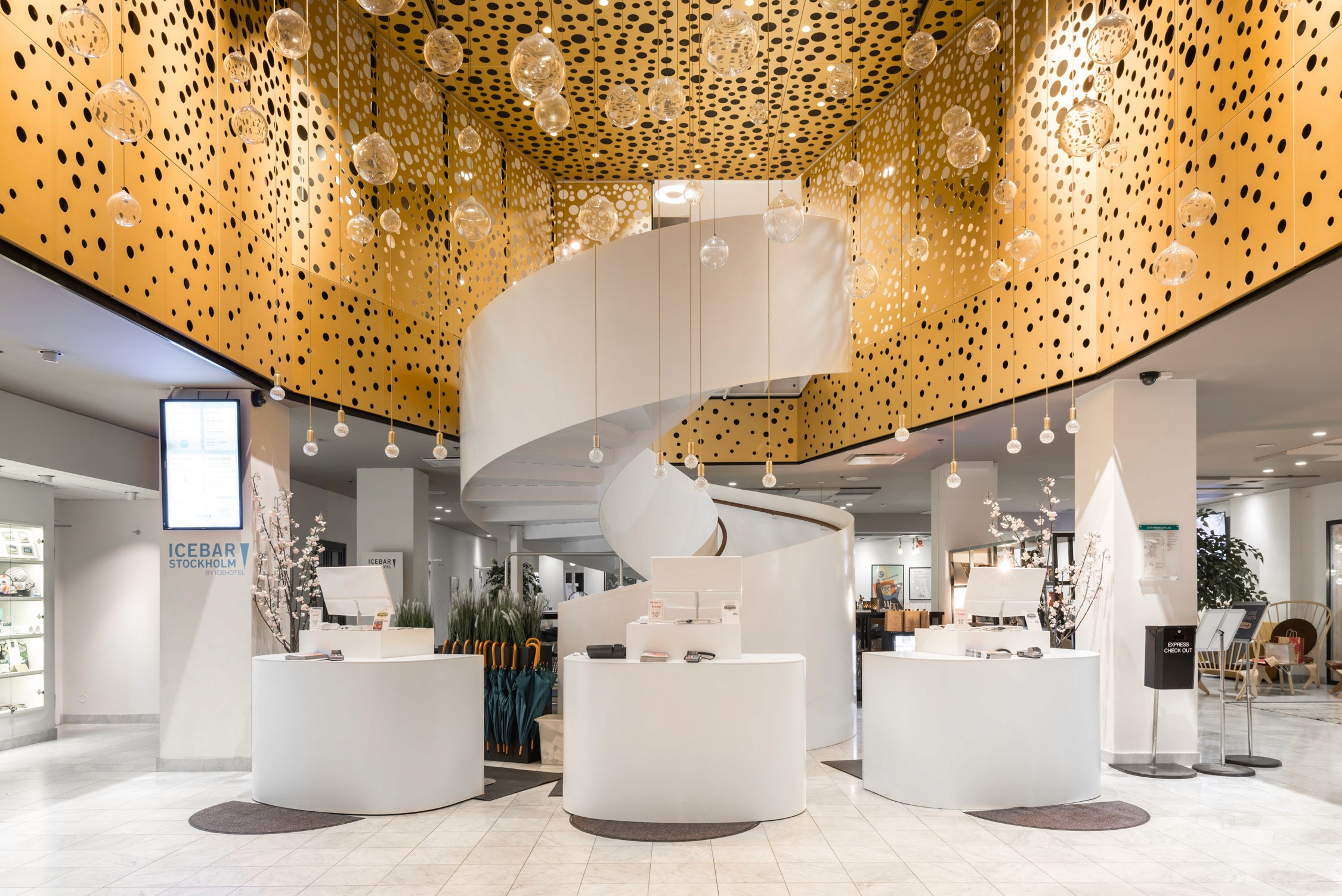 Brightly lit retail space features white counters beneath a striking gold patterned ceiling adorned with hanging glass orbs.