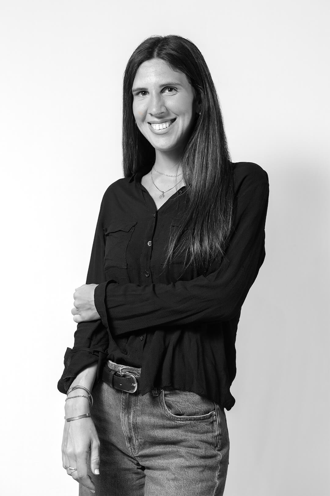 Black and white photo of a smiling woman with long dark hair wearing a black shirt and jeans, standing confidently with a casual pose against a plain background.