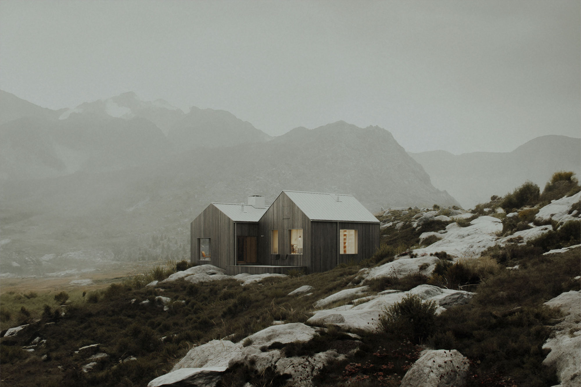 Wooden cabin with warm-lit windows and a metal roof, sitting on a rocky, grassy slope in a misty mountain valley
