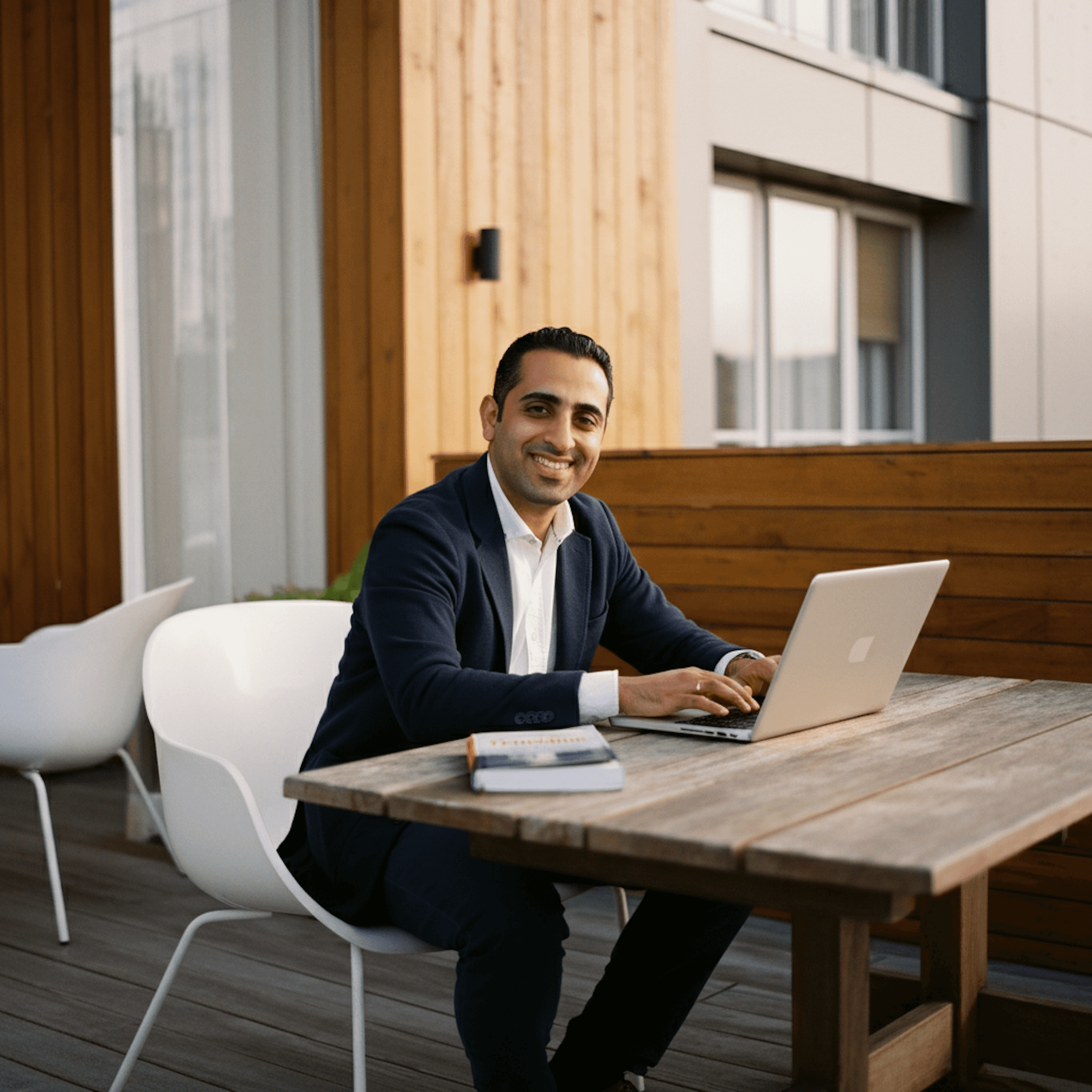 A person in a suit sits at a wooden table, working on a laptop outdoors, smiling at the camera.