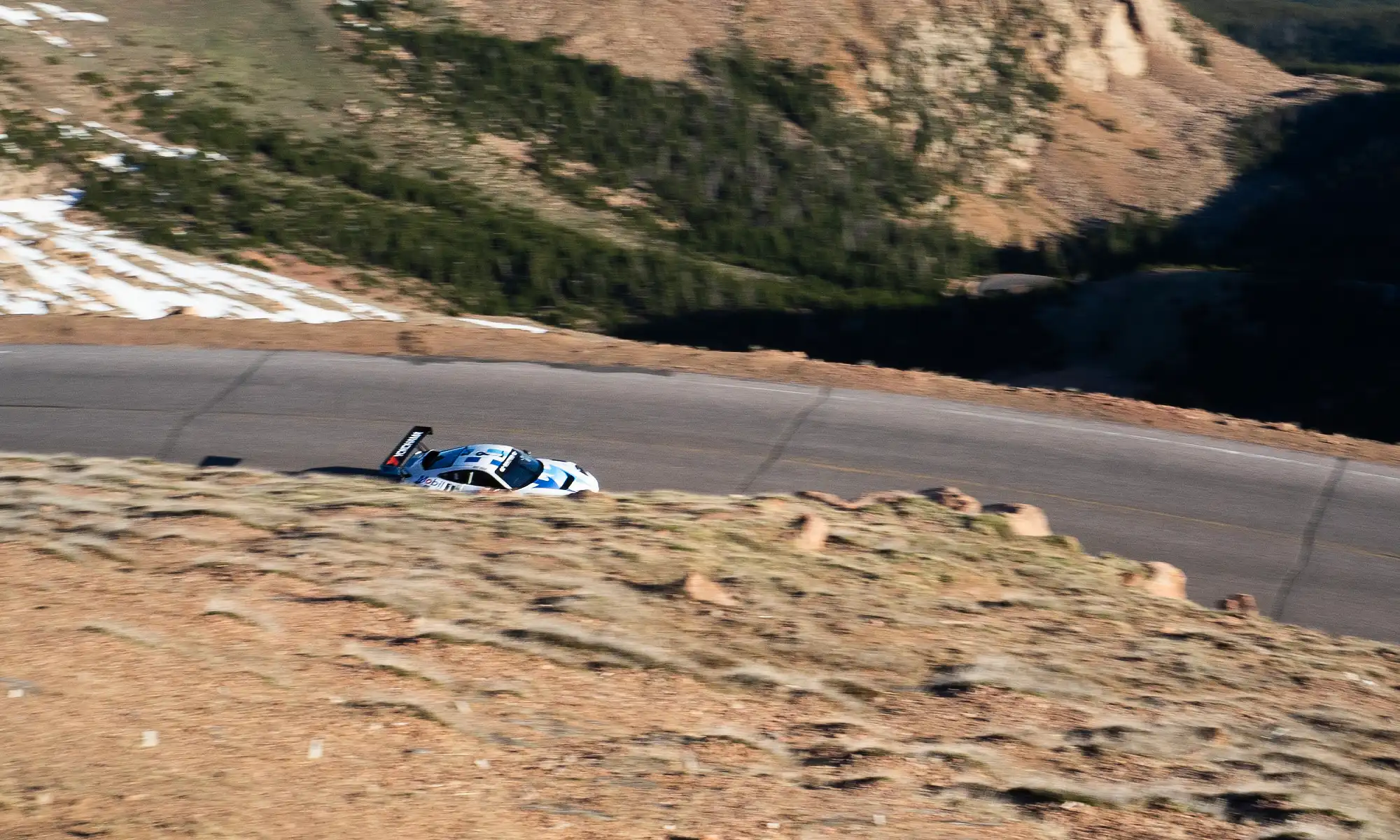 An overhead shot of a white Porsche race car driving on a Pikes Peak mountain road, with snow on the hills around it.