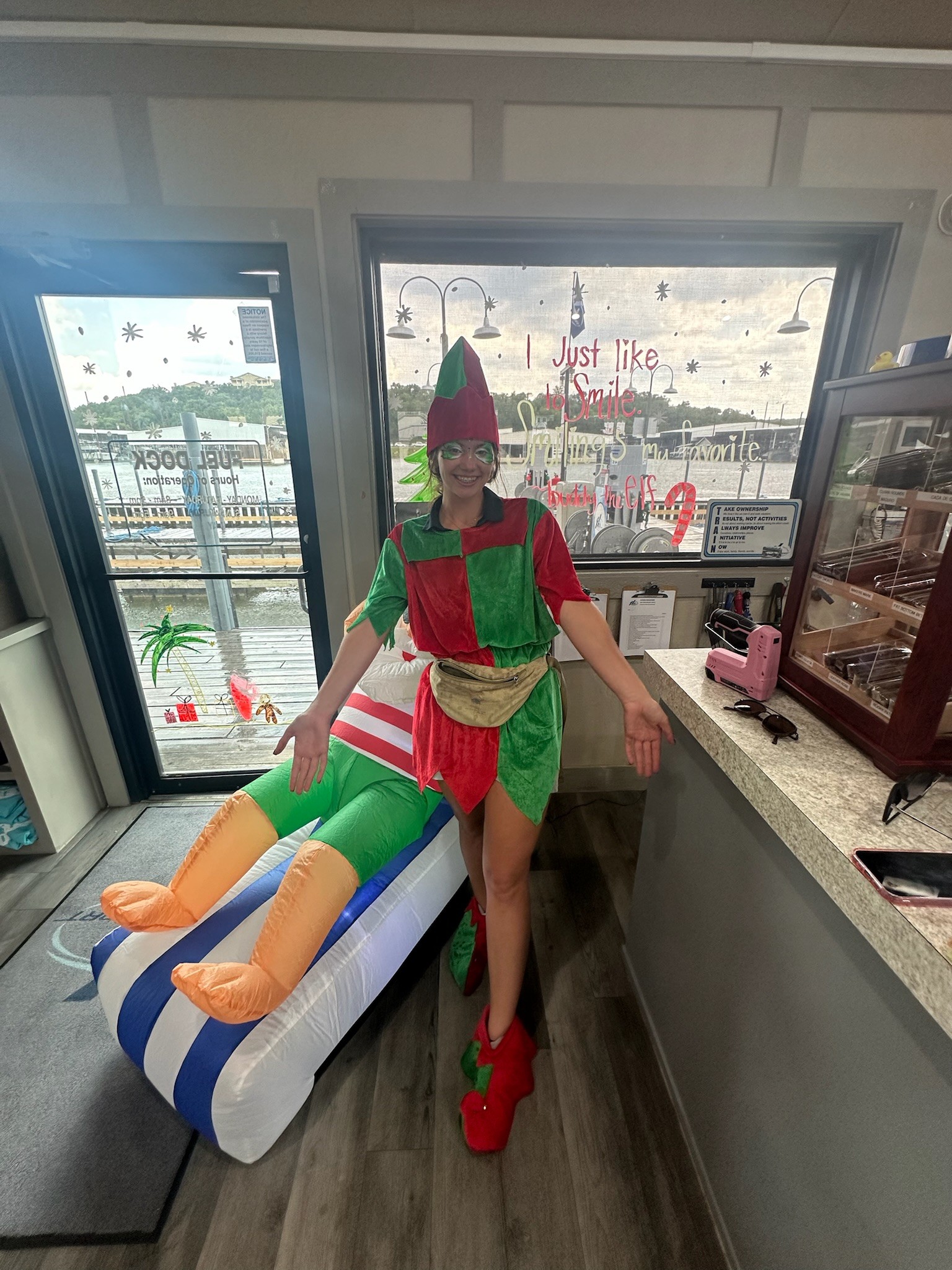 A person dressed in a festive elf costume, complete with a red and green hat and pointed shoes, stands cheerfully beside an inflatable holiday decoration in an office setting, with a window displaying Christmas-themed decals in the background.