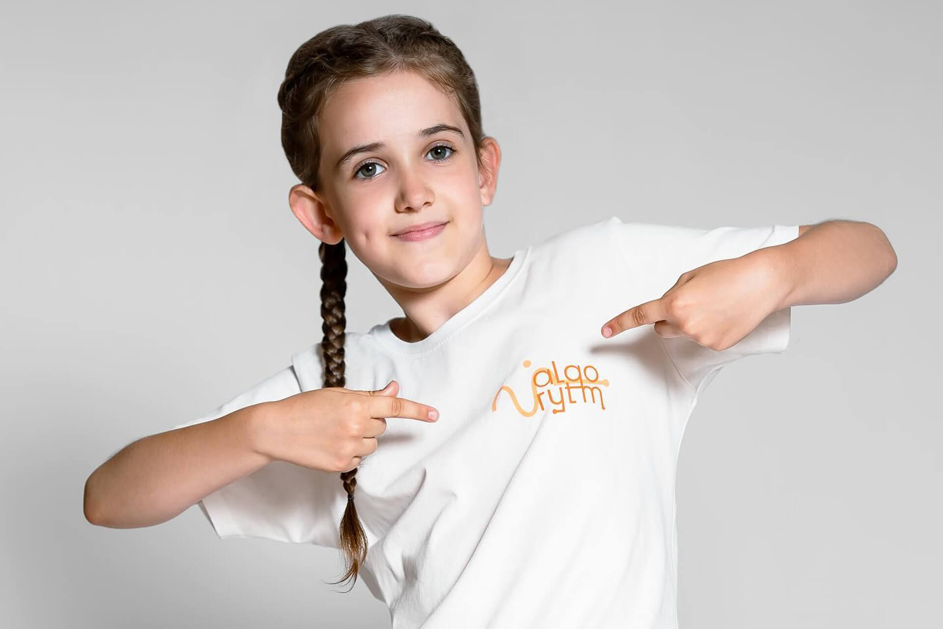 Dziecko z warkoczem w białej koszulce radośnie wskazuje palcami na logo szkoły tańca algoRYTM na szarym tle