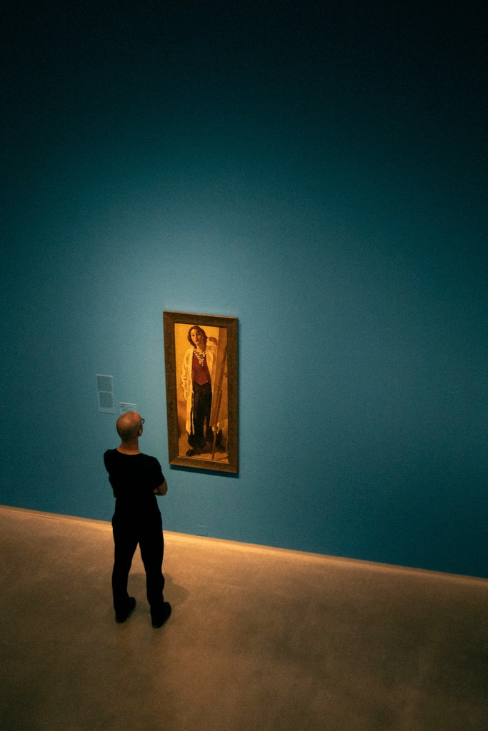 A man stands thoughtfully in front of a painting in an art gallery with deep blue walls. The painting, featuring a figure in traditional attire, is illuminated by focused lighting, creating a contemplative and serene atmosphere.