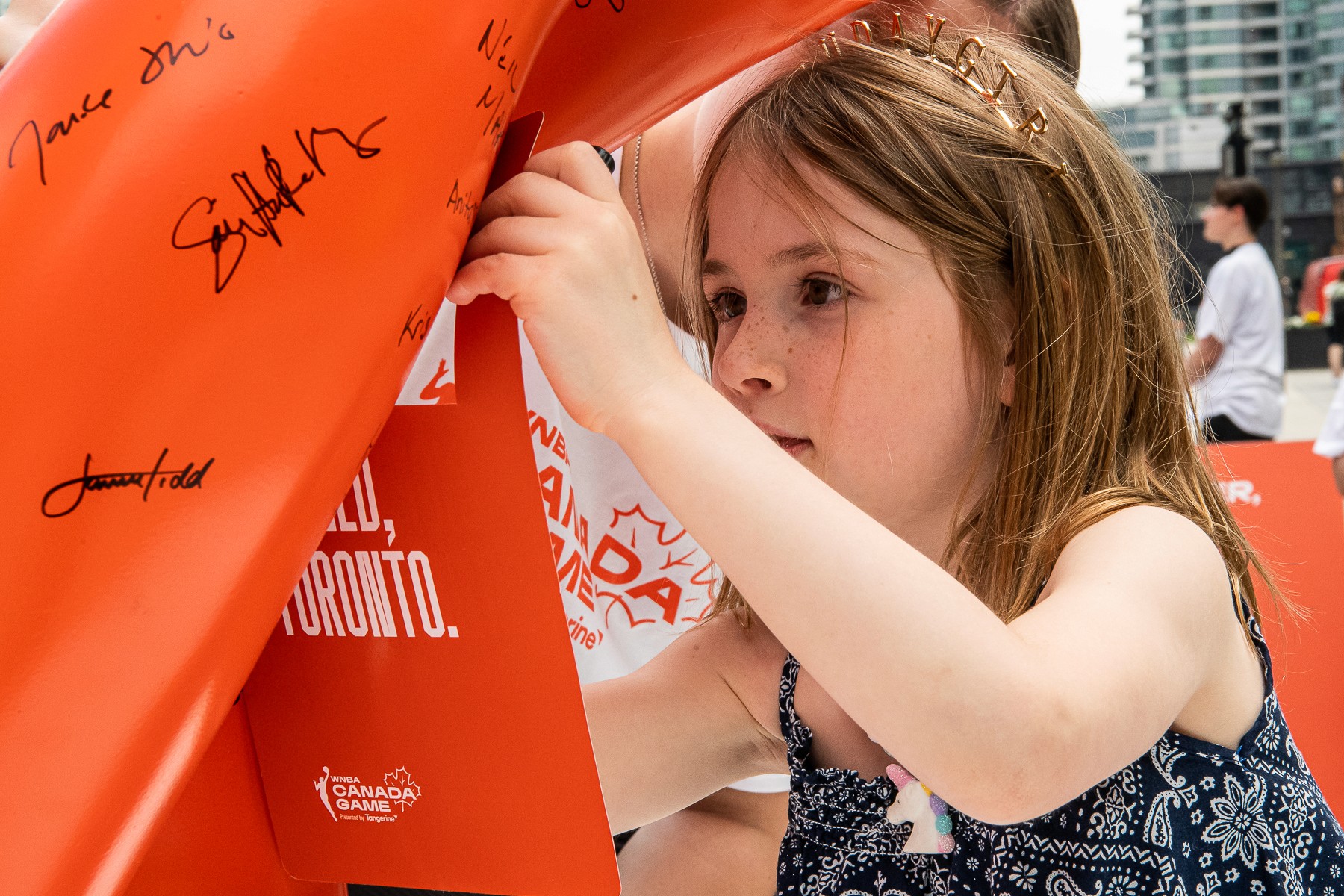 A child in a swimsuit playfully jumps with a water spray in a backyard, surrounded by toys and greenery.