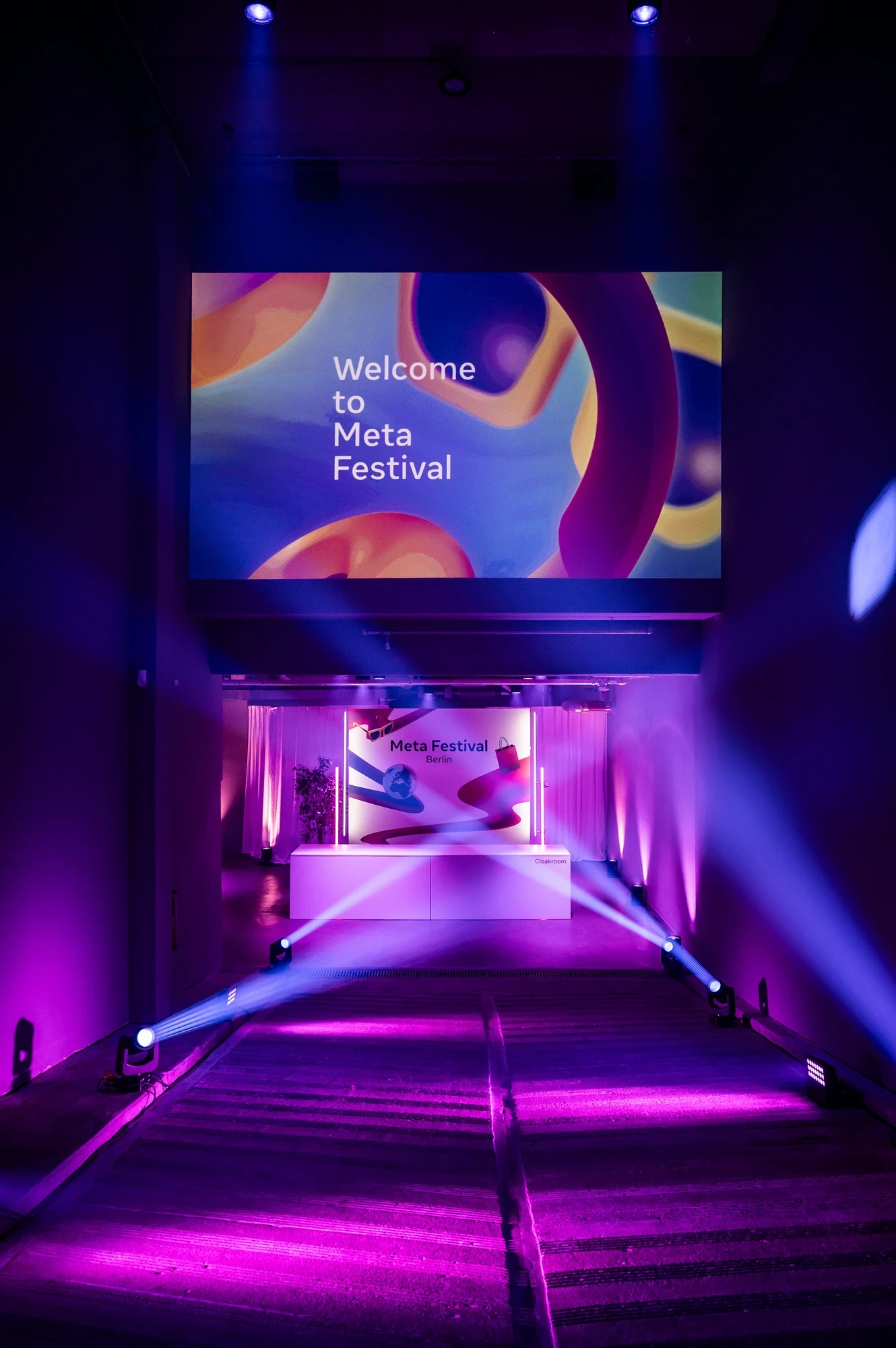 Entrance area of the Meta Festival featuring a large welcome screen above an illuminated staircase, dramatic pink and purple lighting with spotlight beams.