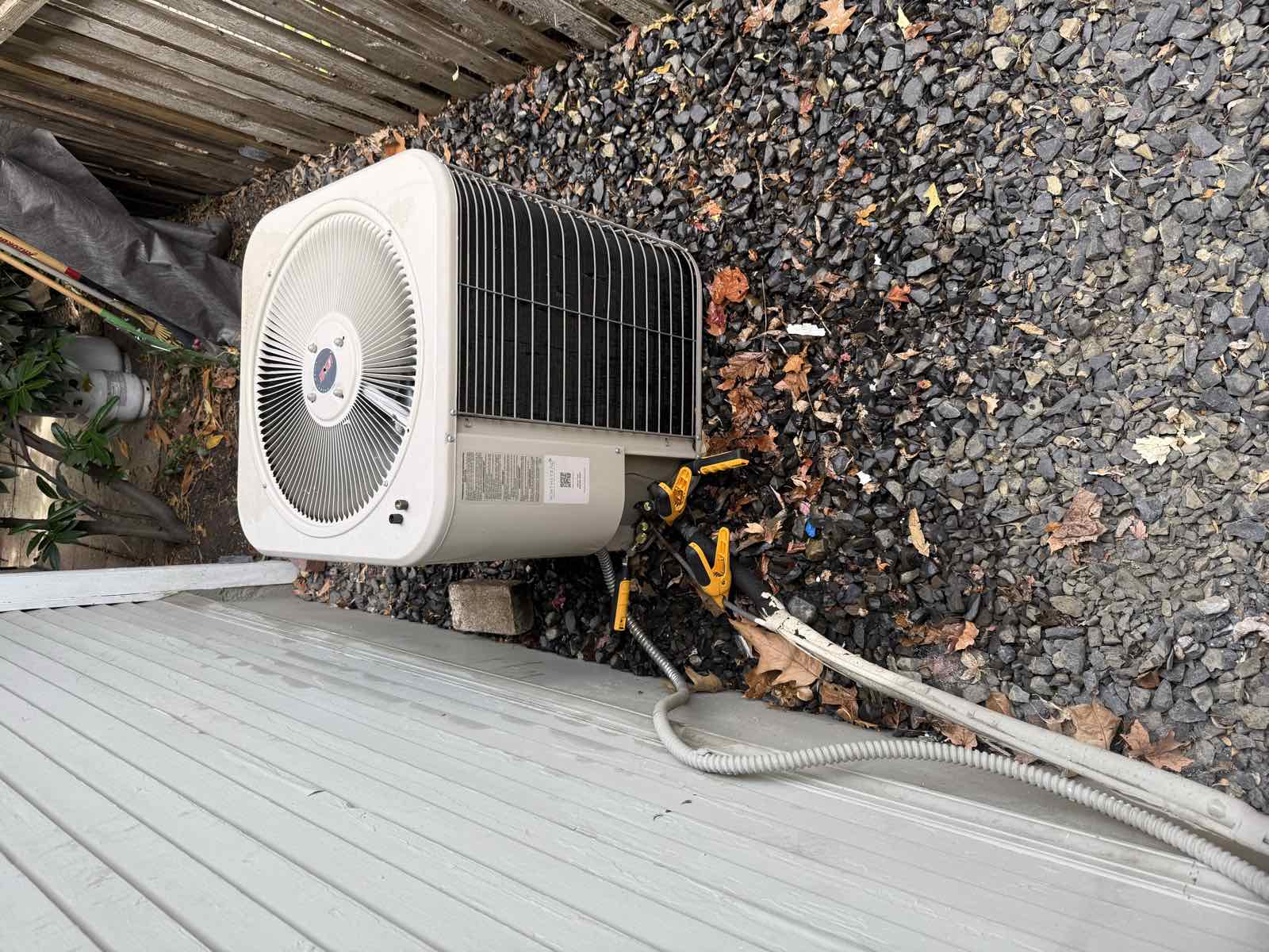 An older residential air conditioning condenser beside a house, on a gravel bed against a wooden fence.