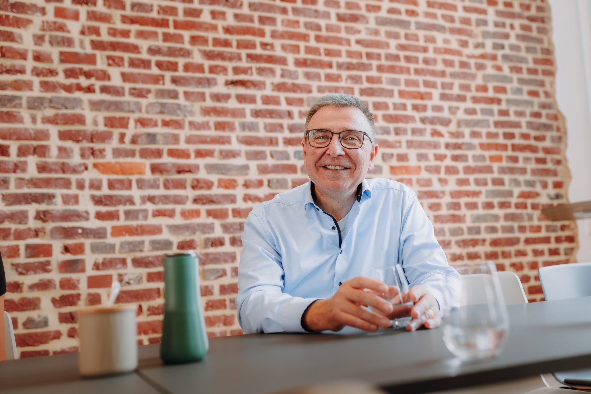 Ein Geschäftsführer älteren Alters mit grauen Haaren und Brille lächelt freundlich in die Kamera. Er sitzt an einem schwarzen Tisch vor roter Ziegelwand, trägt ein hellblaues Hemd und hält ein Glas Wasser in der Hand.