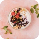 A bowl of yogurt topped with blackberries, strawberries, and granola, surrounded by fresh mint leaves on a pink backdrop.