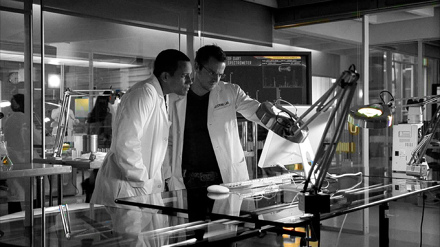 Black and white photo of a forensics lab. 2 male researchers in close-up, dressed in lab white overall discussing over what they see on a desktop computer monitor.