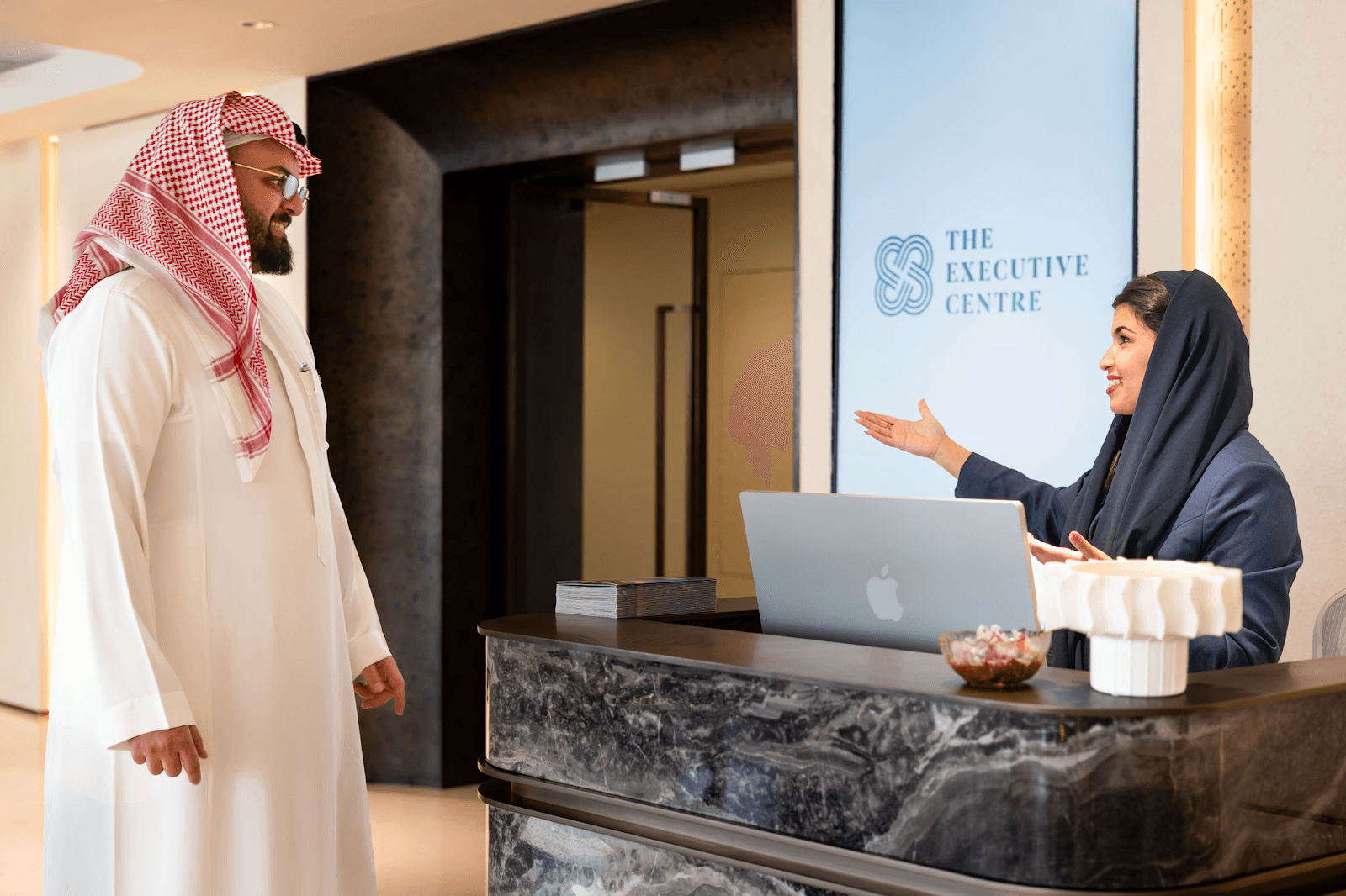 صورة لموظفة استقبال خلف كونتر رخامي تتحدث مع عميل عن خدمات حجز الاسم التجاري.