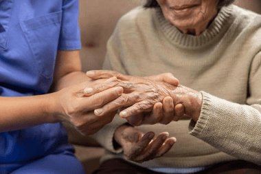 Elderly person's hands gently held by a caregiver in blue scrubs indoors.