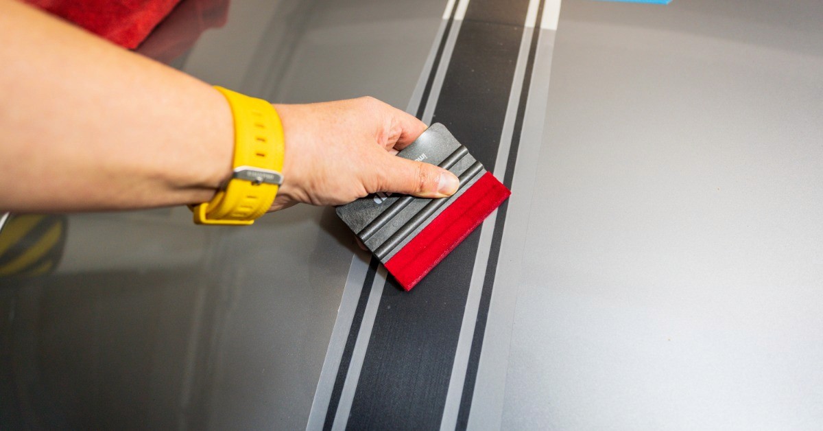 A person is using a red and black squeegee to secure a black stripe vinyl decal across the hood of a gray car.