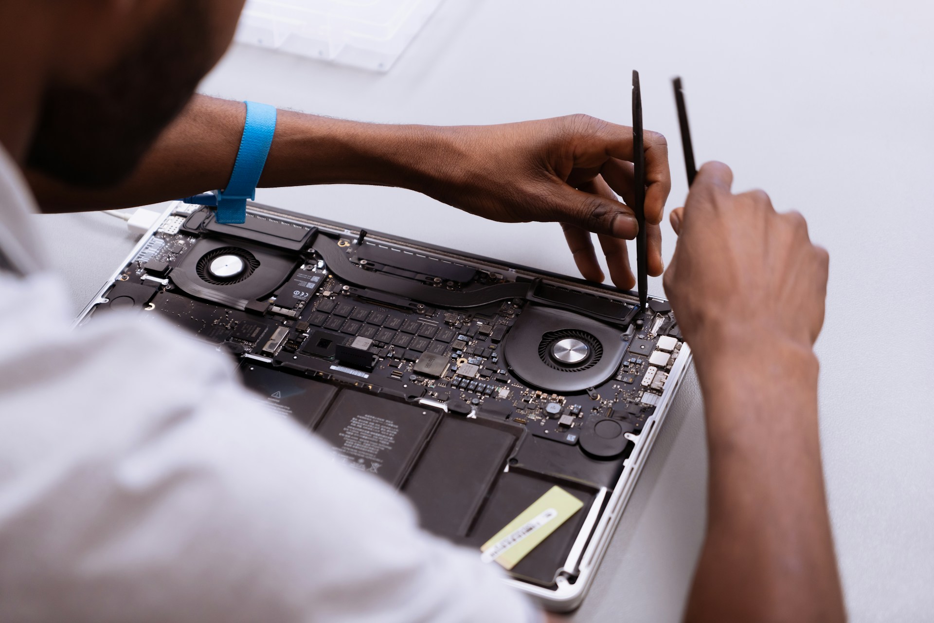 A person's hands are using a pair of tweezers to work on the exposed circuit board and internal components of an open laptop, indicating repair or maintenance.
