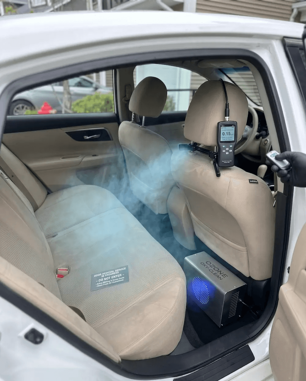 A high-angle, realistic view of a white sedan's rear interior during a professional odor removal service. An industrial stainless steel ozone generator sits on the black floor mat, emitting a visible fine mist into the beige cabin. A digital air quality monitor is clipped to the front passenger headrest, displaying a reading of 0.15, while a hand in a black nitrile glove holds a remote control in the foreground. A professional "Do Not Enter" service placard is placed neatly on the rear bench seat.