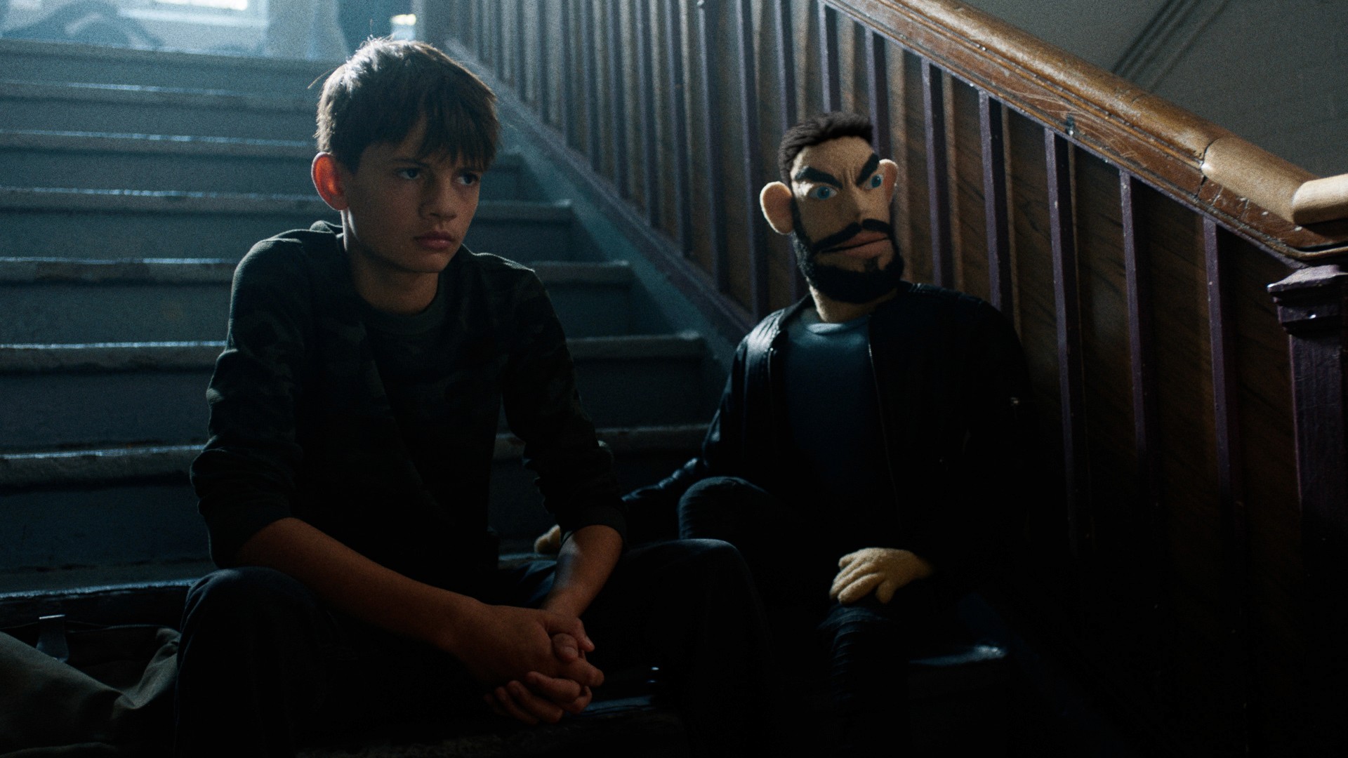Boy sitting pensively on dimly lit stairs next to a puppet with dark hair and beard. The scene appears tense as both figures have a serious and expression on their face
