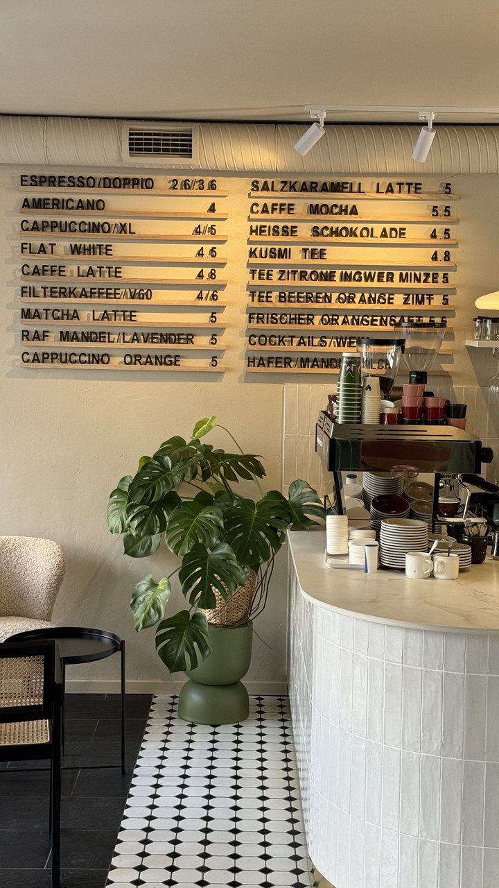A cozy café interior featuring a menu board with various coffee options, a green potted plant on a black-and-white checkered floor, and a curved, white counter displaying coffee equipment and mugs.