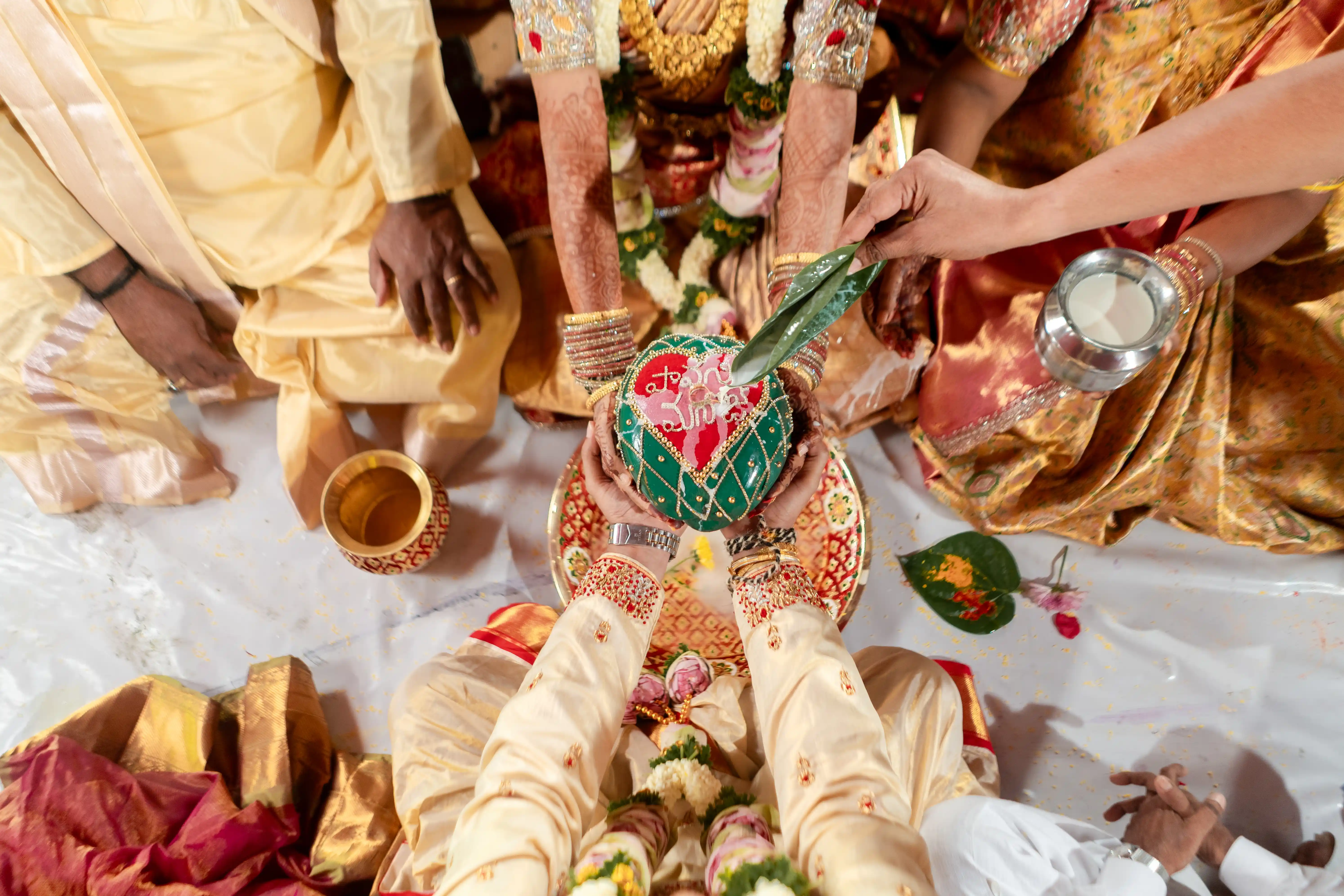 Top-down view of a traditional wedding ritual where the bride and groom hold a decorative coconut adorned with symbols, surrounded by family members, skillfully captured by Out of The Blues Fine Art Wedding Photography in Hyderabad.