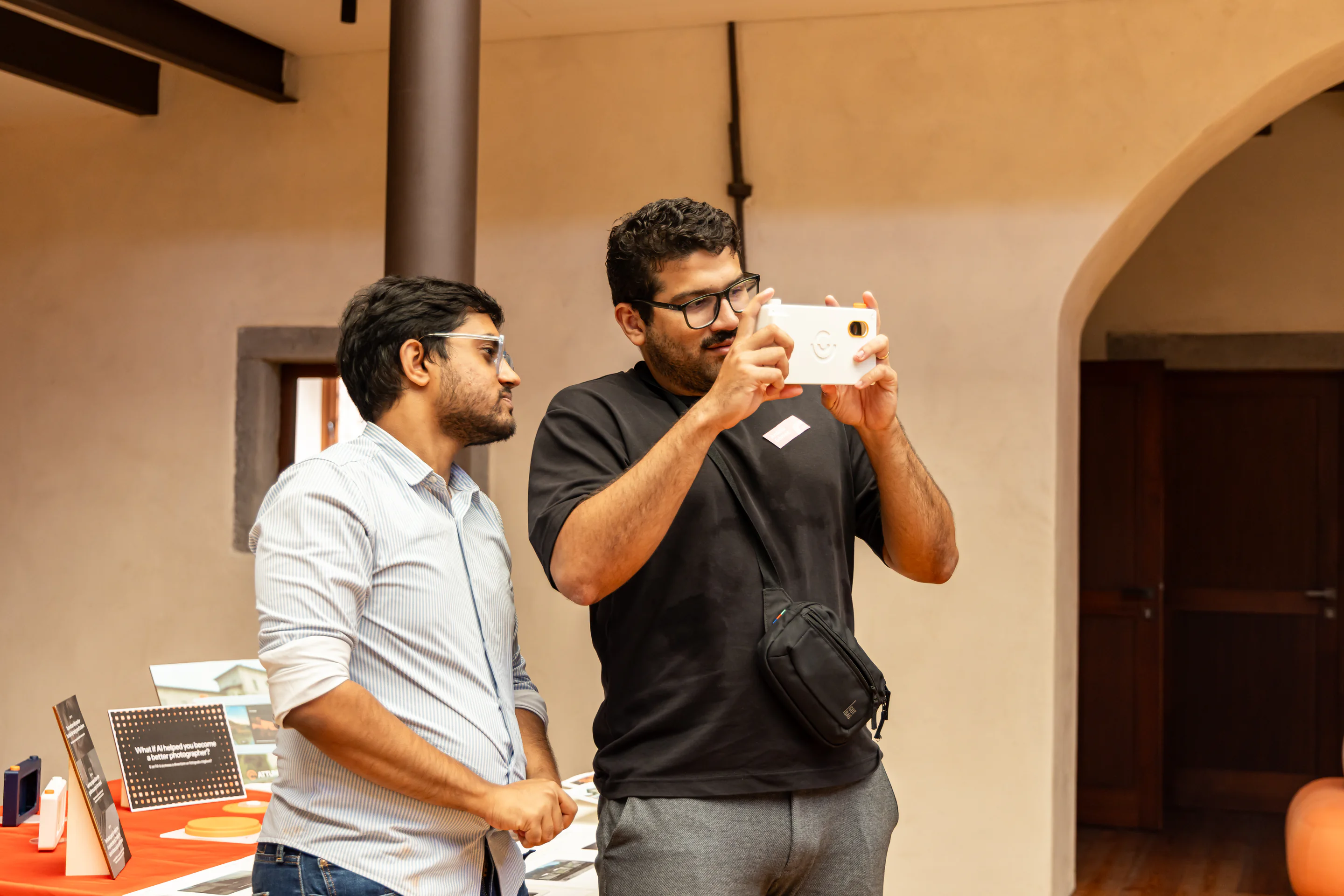 One person holds a white camera prototype in shooting position while another stands nearby.