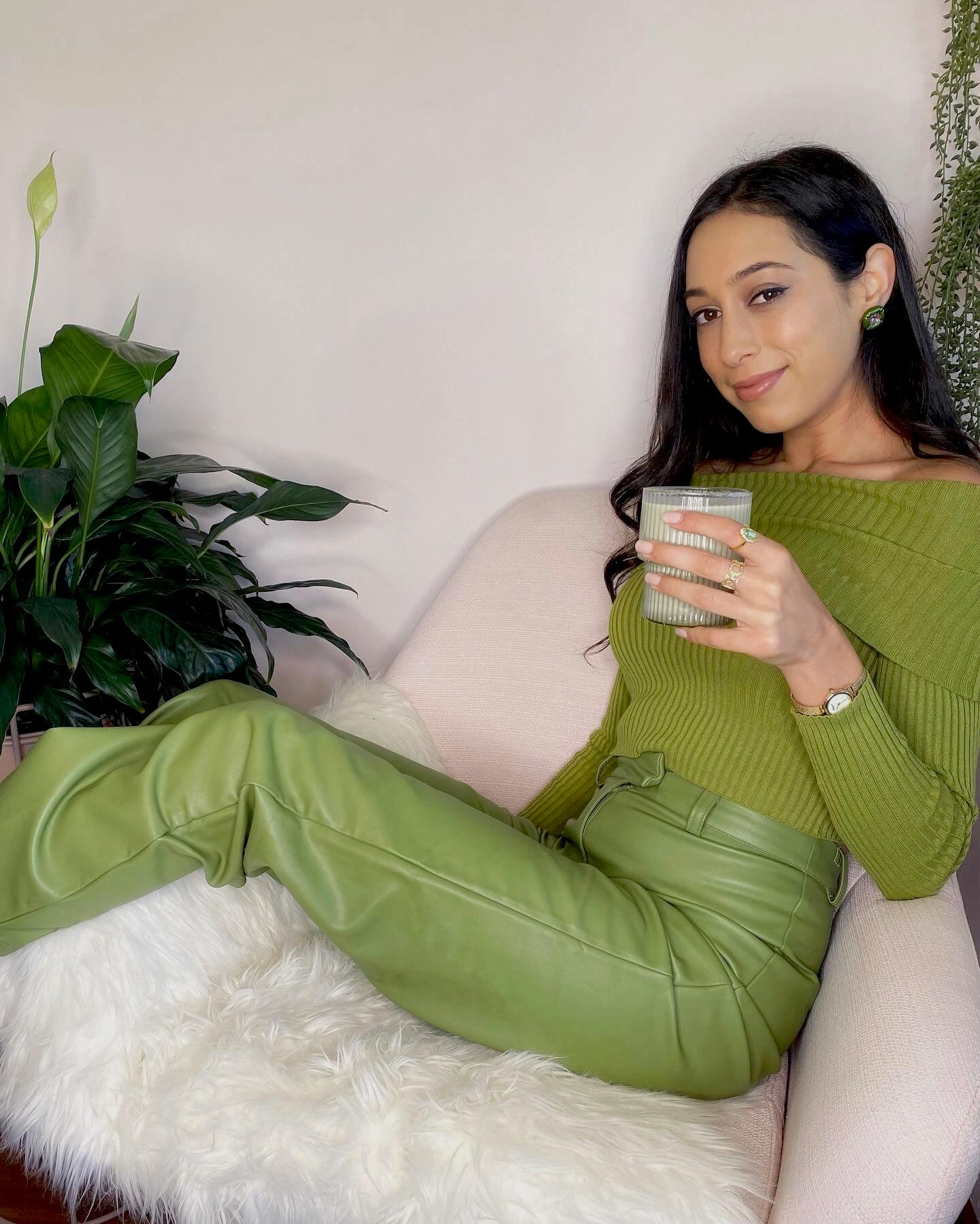 Girl wearing all green, setting on a pink sofa chair, holding a matcha latte, against a pink painted wall with plants in the background