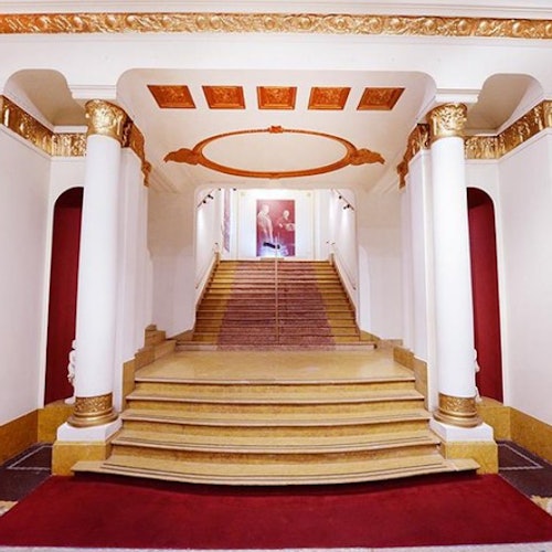 Elegant staircase with white and gold columns, red carpet, and a decorative ceiling leading to a portrait at the top.