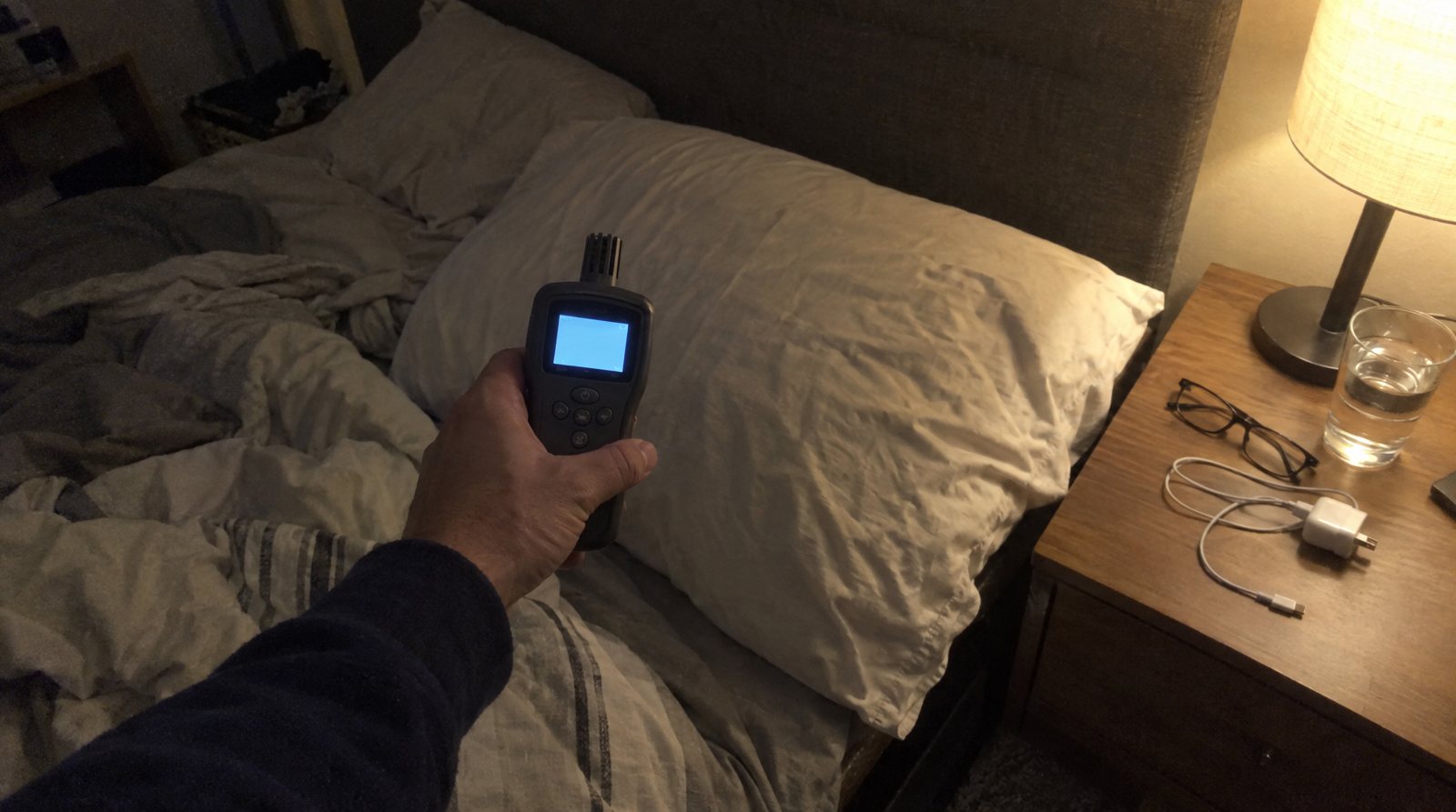 A hand holding a particle counter at pillow height beside a bed at night.