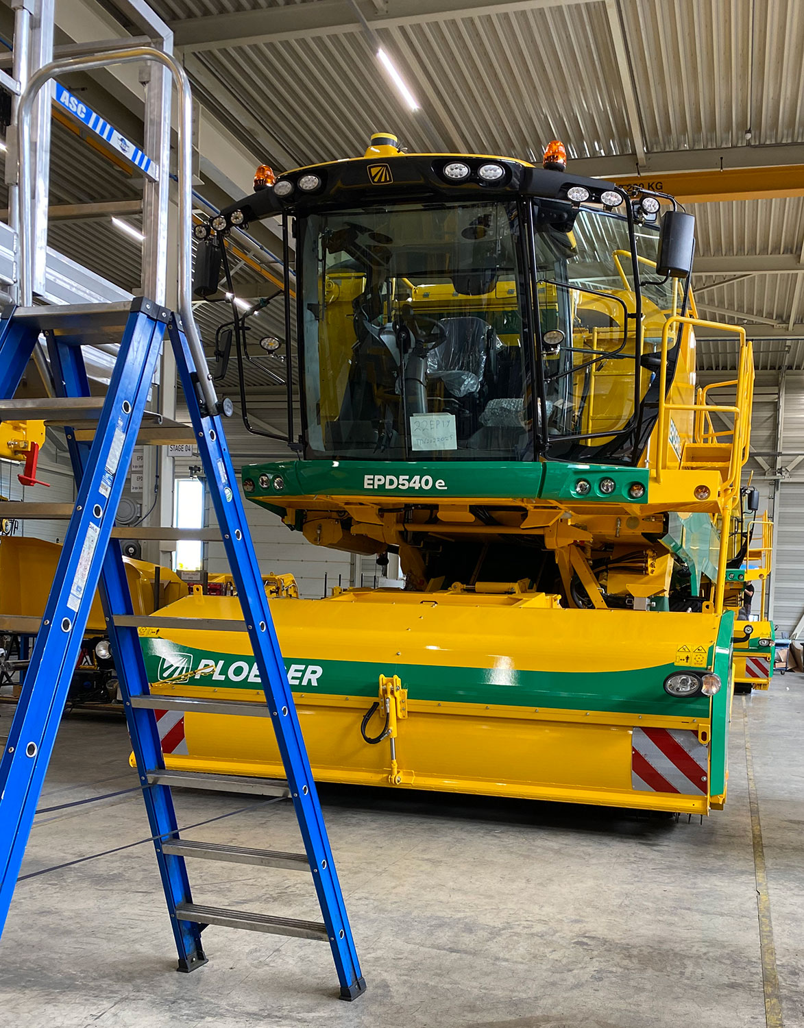 Front view of a bright yellow and green Ploeger pea harvester. There is a blue ladder standing in front of the harvester.