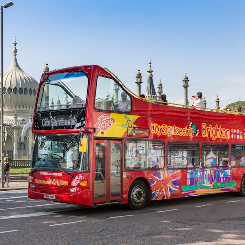 Red double-decker sightseeing bus in Brighton with tourists on the top deck, passing by a building with domed architecture.