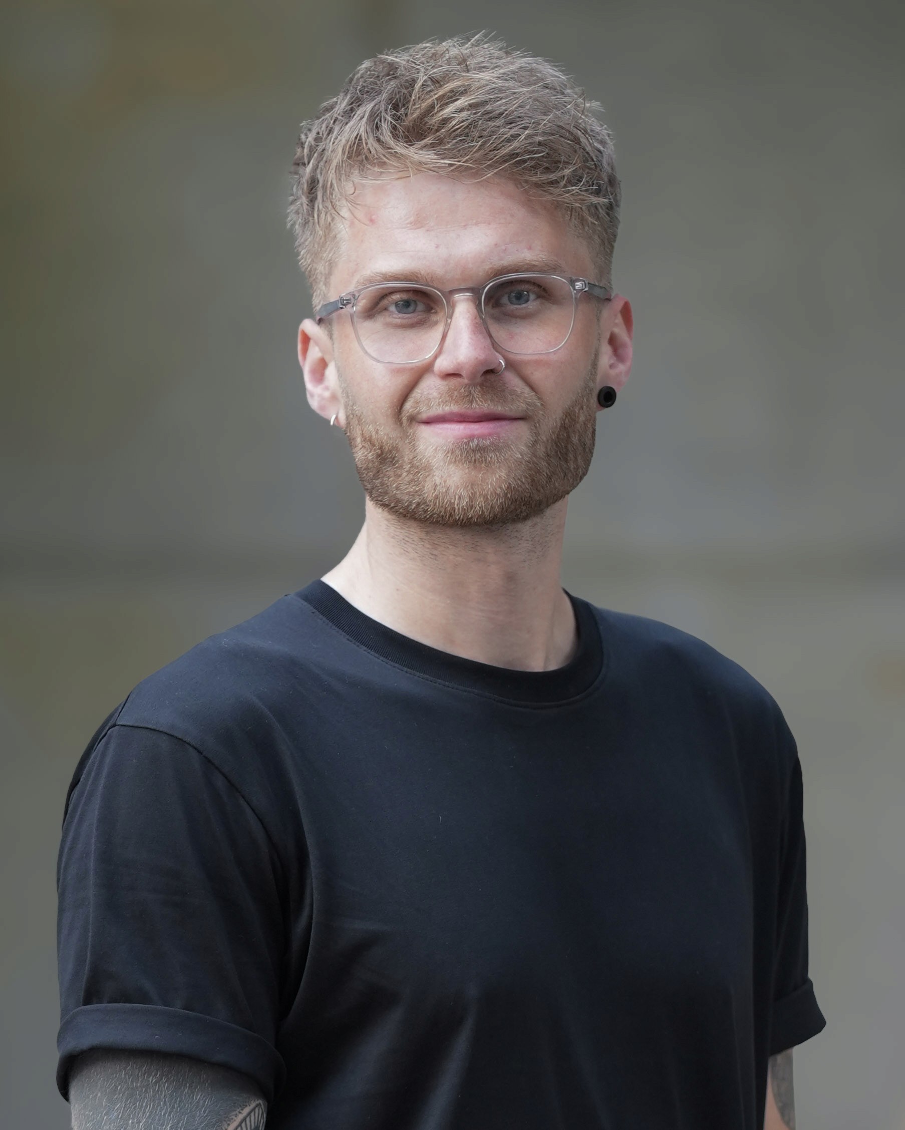 A smiling man with short hair stands with arms crossed, wearing a dark shirt against a neutral background.