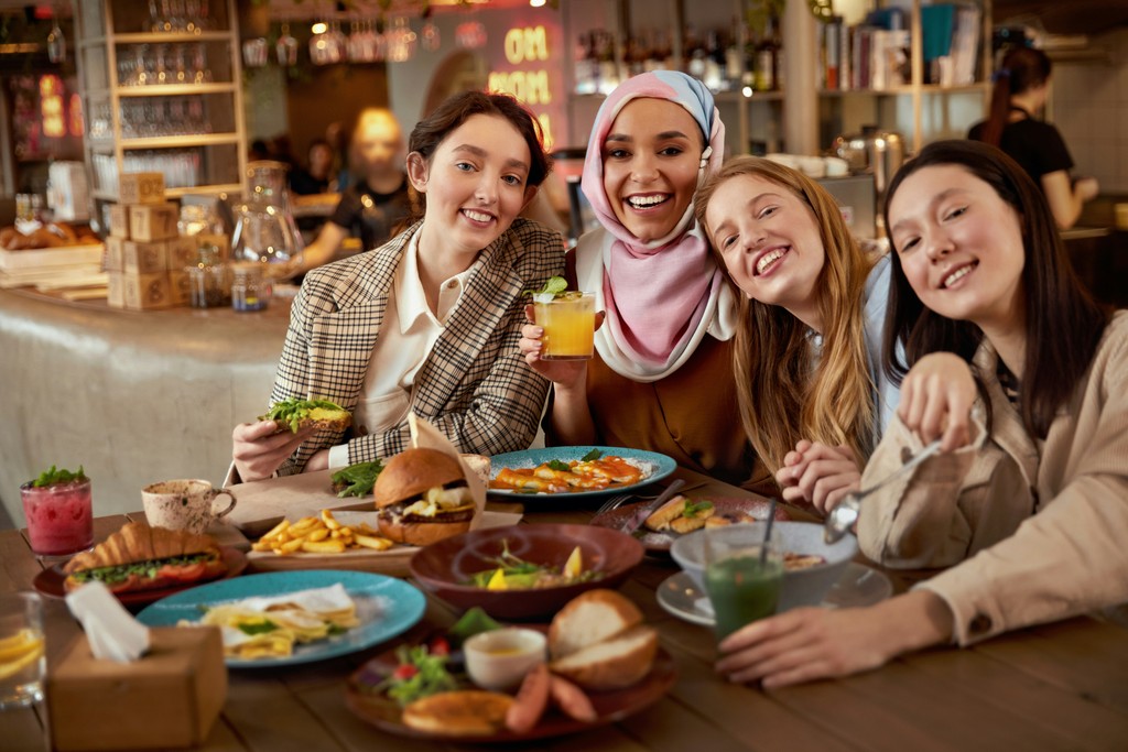 A group of four diverse women enjoy a meal together in a cozy, well-lit restaurant. They are smiling and laughing, with a table full of various delicious dishes and drinks in front of them. The atmosphere is warm and lively, reflecting friendship and shared moments.