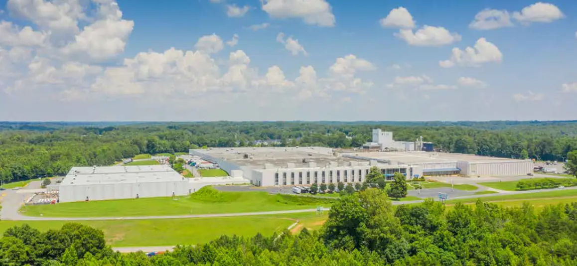 Aerial view of a large industrial facility surrounded by lush greenery under a blue sky with fluffy clouds, conveying a sense of scale and tranquility.