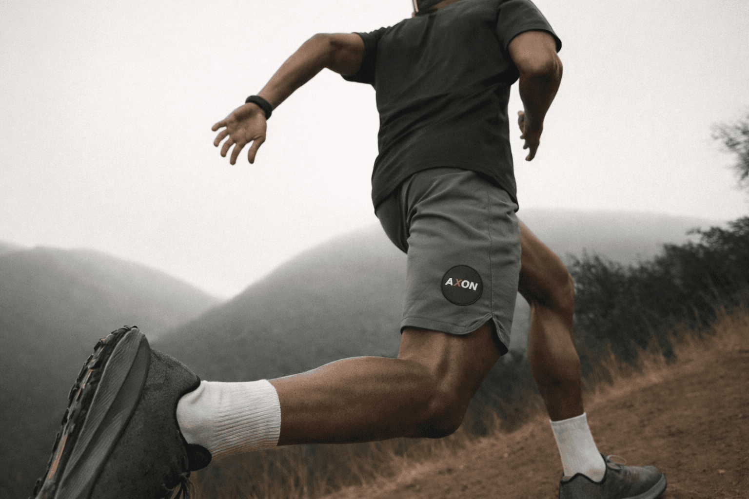 Athletic man in a sleeveless black outfit posing confidently outdoors under a bright blue sky, with a close-up of navy and salmon-colored men’s running shoes in the foreground.