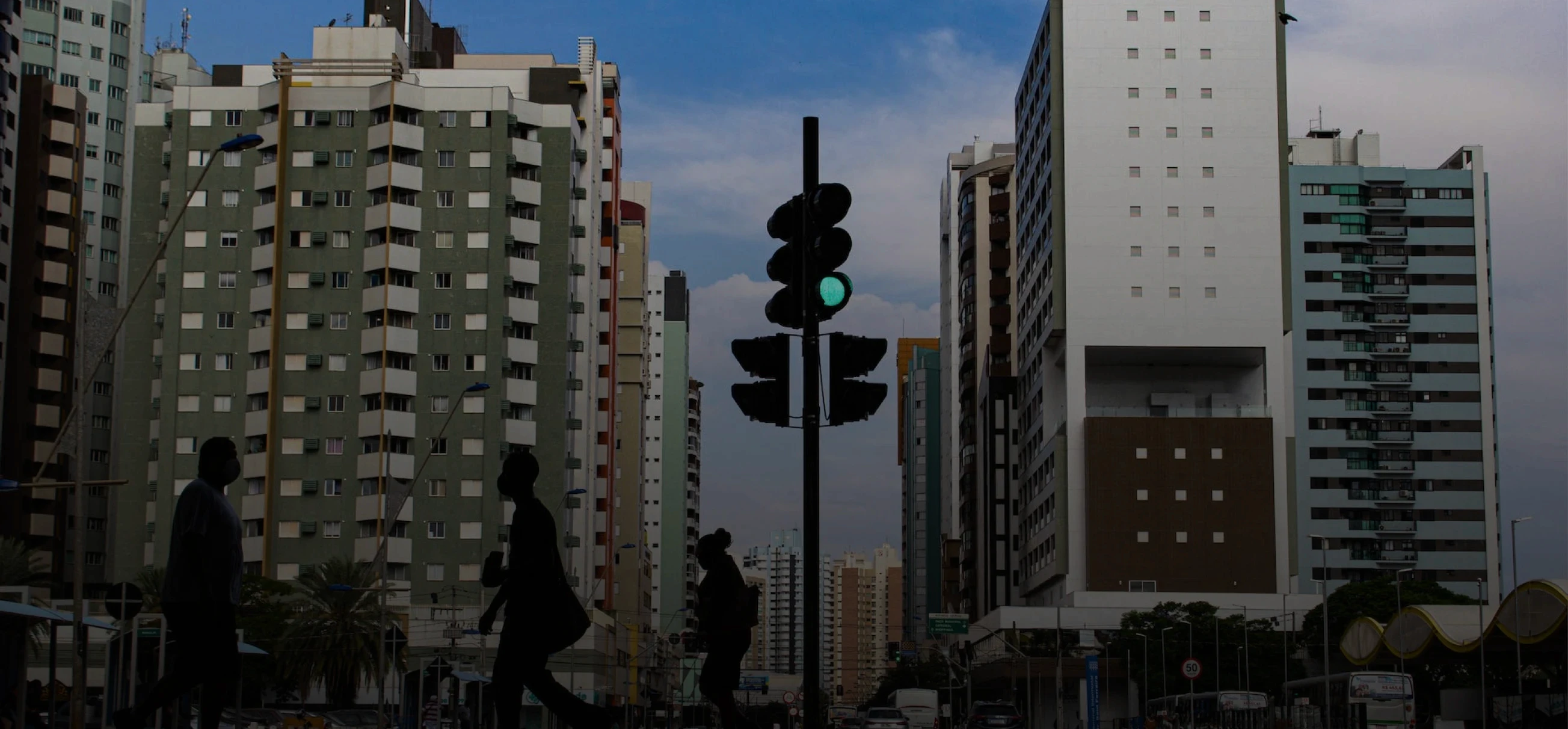 imagem de tráfego urbano, com algumas pessoas atravessando a rua, carros passando aos lados e prédios no fundo da imagem