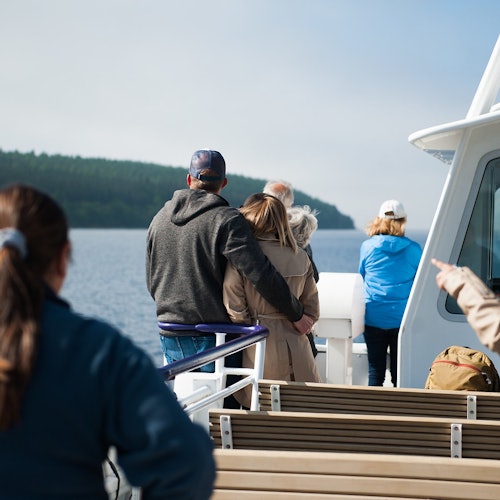 Personas de pie y sentadas en un barco, con un lago y colinas cubiertas de árboles al fondo.