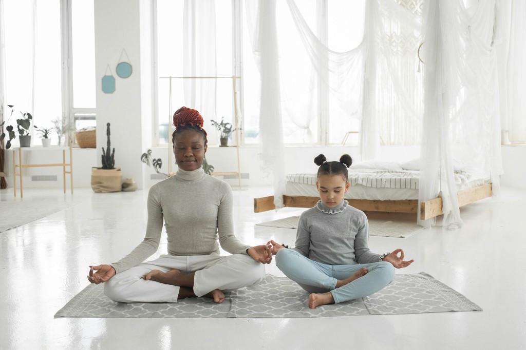 A woman and a young girl meditating together in a serene, bright room with minimalist decor, sitting cross-legged on yoga mats and practicing mindfulness.