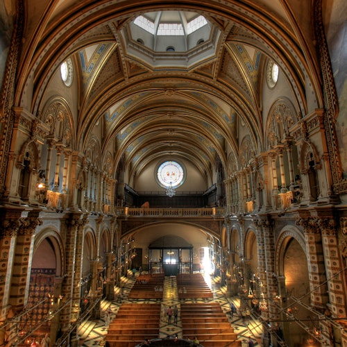 Interior of an ornate church with arched ceilings, a large circular stained-glass window, and rows of wooden pews in symmetry.